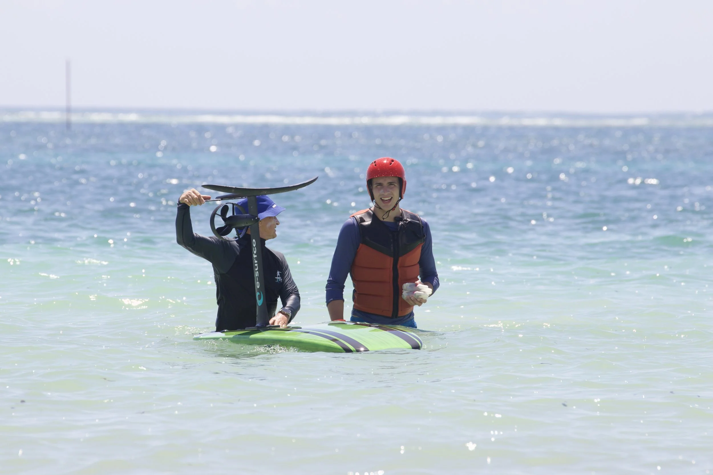 Two people in the water, one holding a paddle and the other smiling and holding a helmet, at the beach with the ocean in the background.