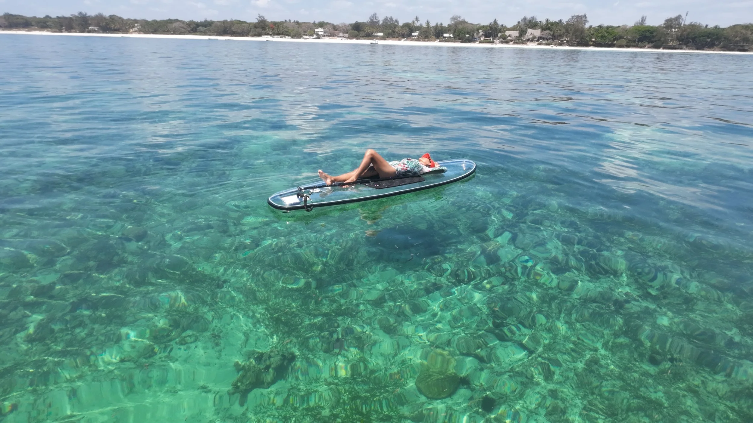 A person lying on a paddleboard floating on clear, turquoise water with underwater rocks visible. In the background, there is a shoreline with trees and houses.