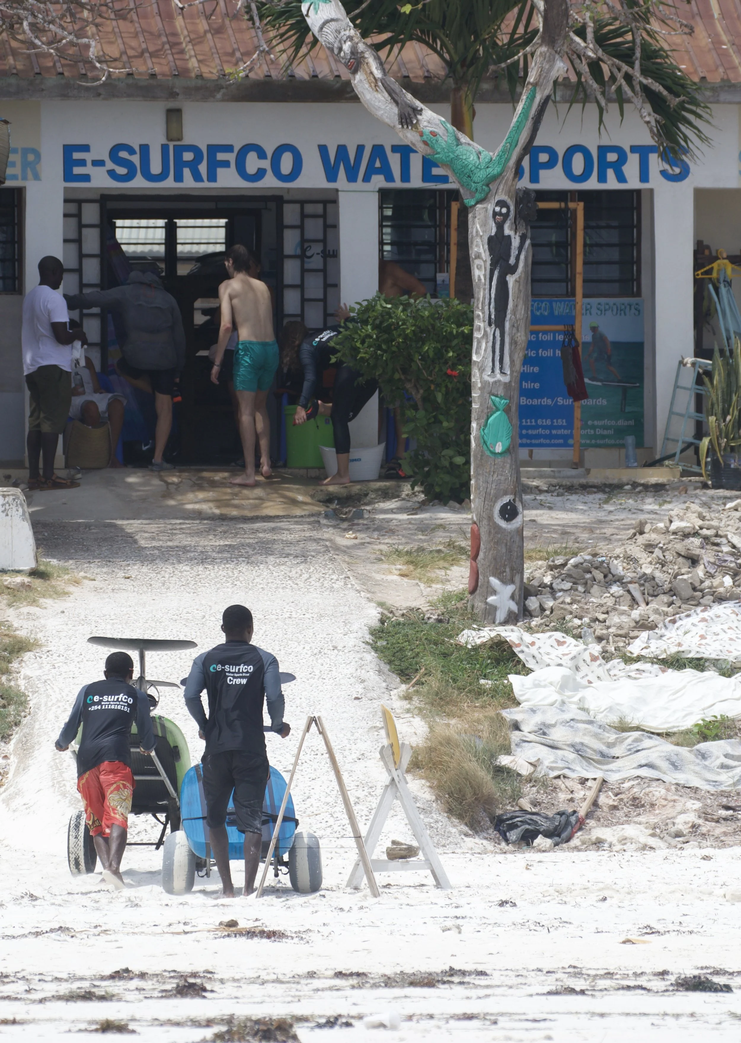 A group of people gather outside a water sports shop with a sign reading 'E-SURFCO WATER SPORTS.' Two crew members are walking towards the beach, pushing a small, blue watercraft. Several other individuals are near the shop entrance preparing equipme