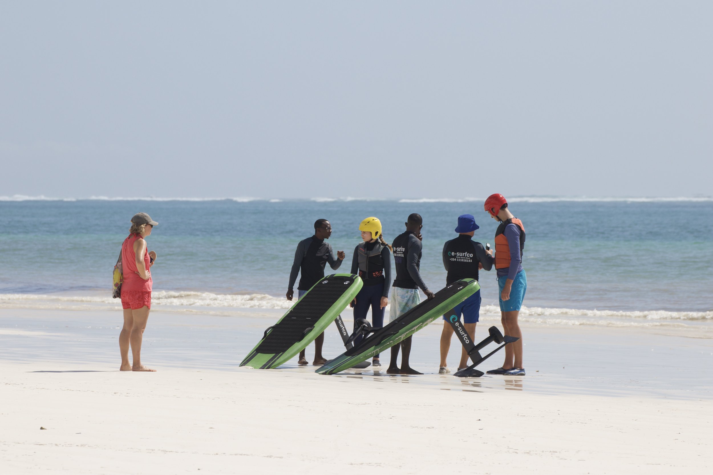 Group of people on a beach, some wearing life vests, helmets, and wetsuits, preparing for water sports near paddleboards and a water scooter.