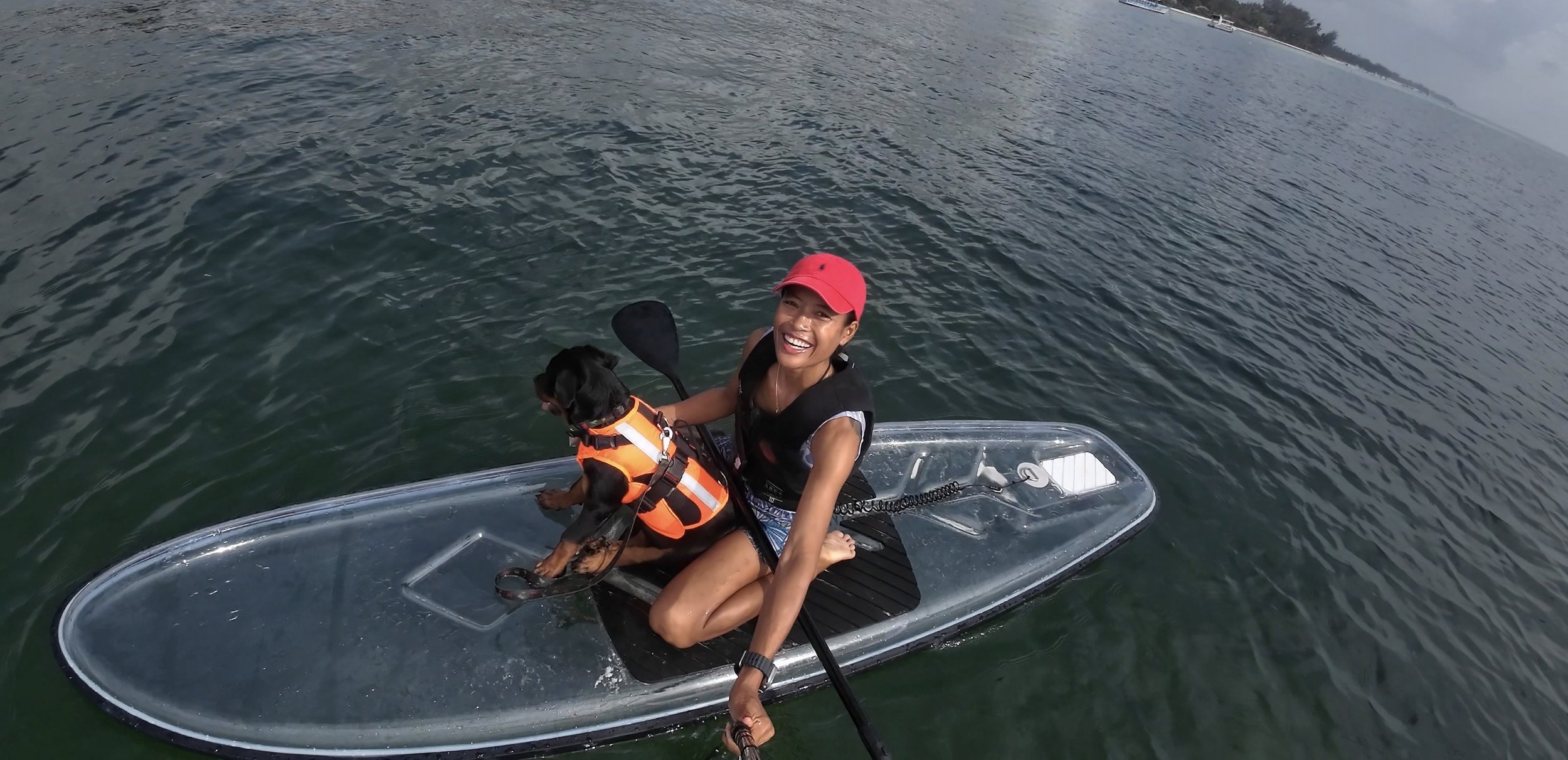 A woman in a red cap smiling on a paddleboard, holding a dog wearing an orange life vest, on a body of water under a cloudy sky.