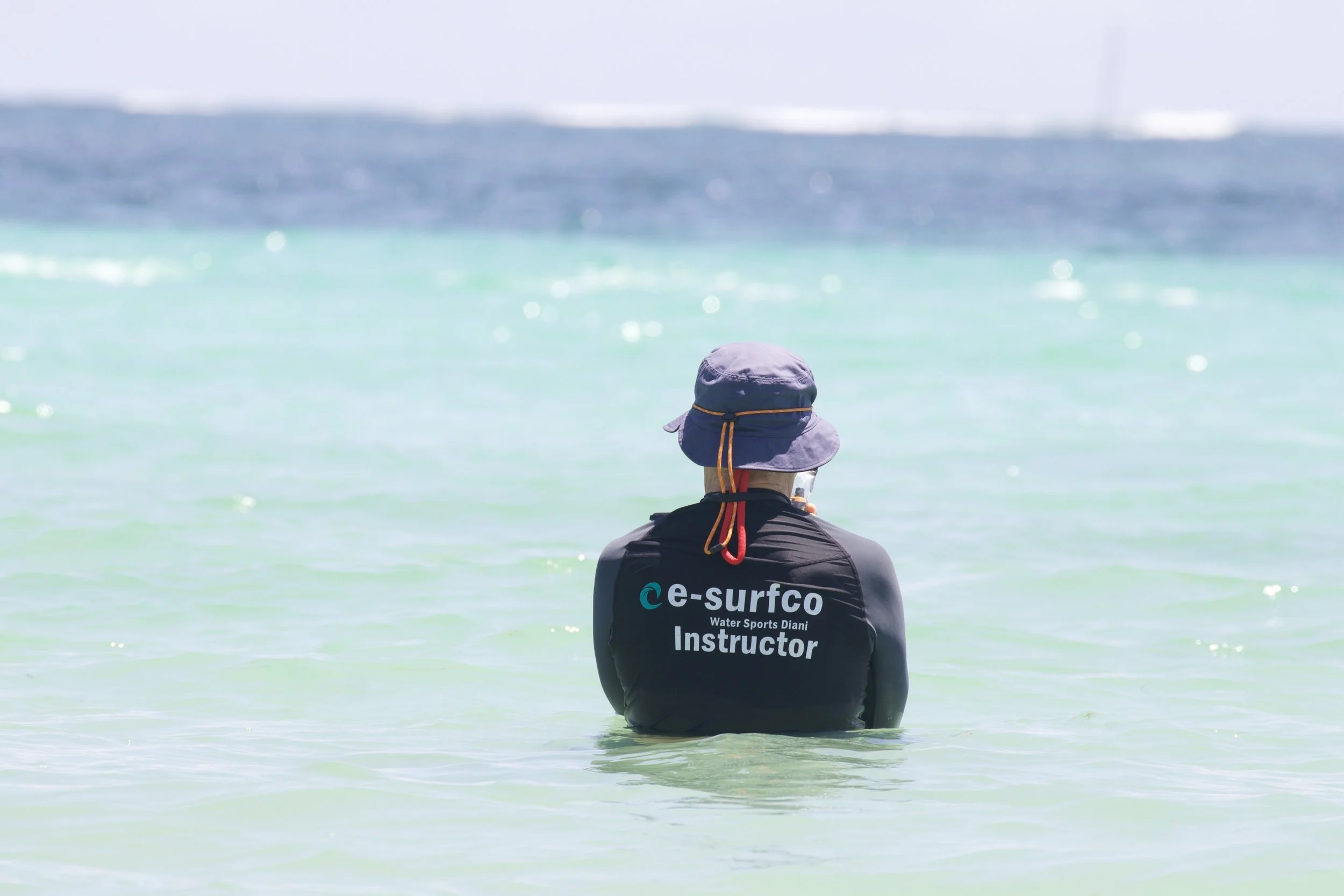 A person wearing a hat and a black wetsuit with 'e-surfco Water Sports Diani Instructor' on the back, standing in shallow clear water with a distant ocean horizon in the background.