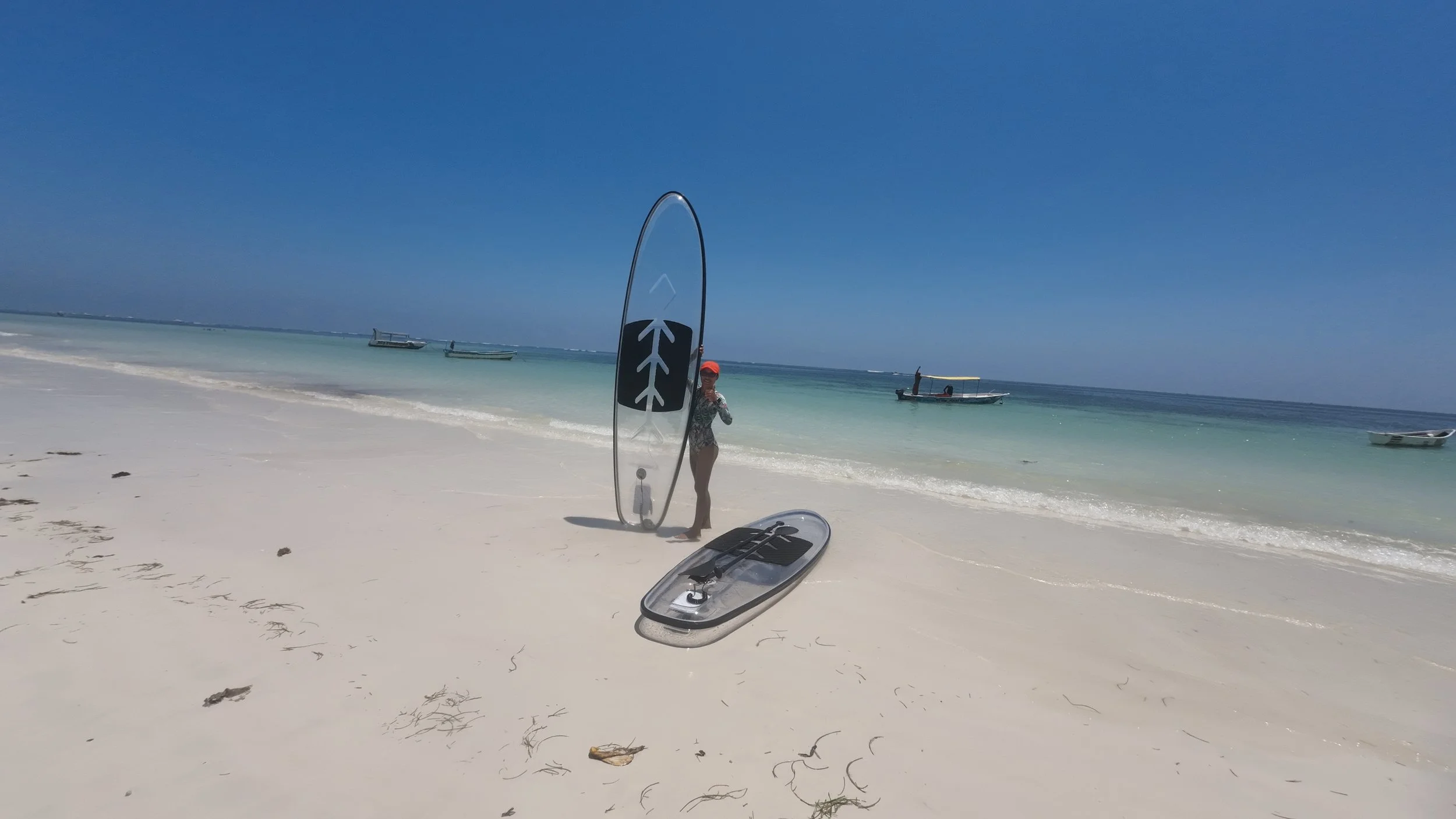 Beach scene with a person holding a surfboard, two other surfboards on the sand, and boats floating on the ocean under a clear blue sky.