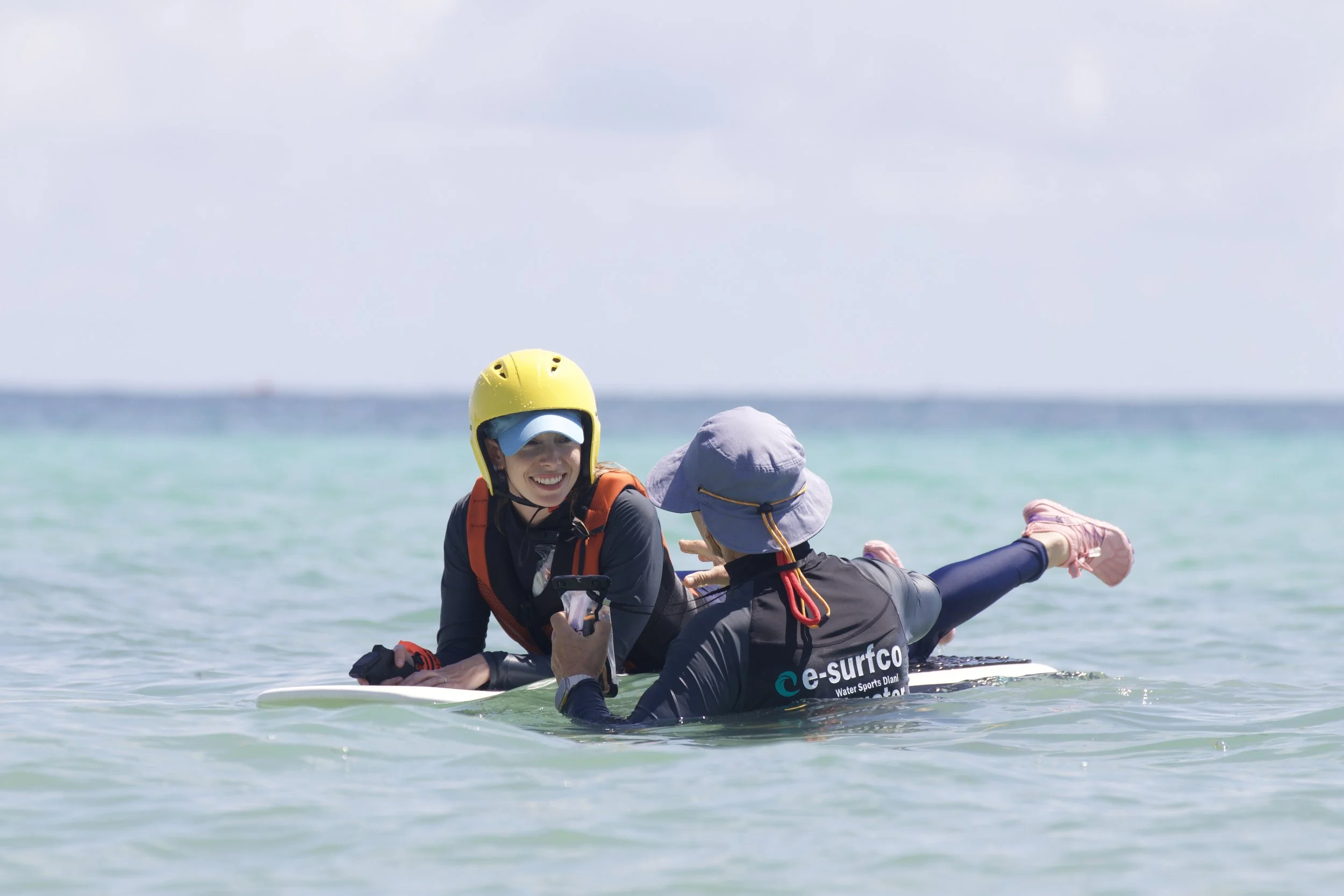 Two women in wetsuits and hats lying on a paddleboard in the water, smiling and talking, with one holding a phone.