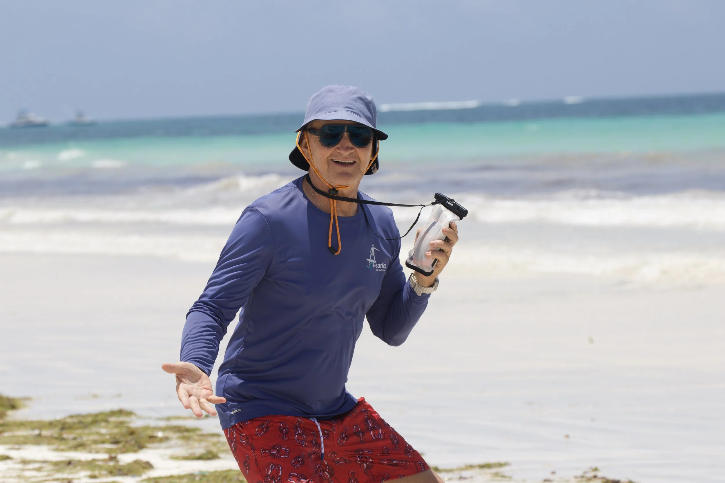A man in a blue long sleeve shirt, red patterned shorts, and a wide hat is smiling and gesturing with his hand on a sunny beach, holding a water bottle and wearing sunglasses.