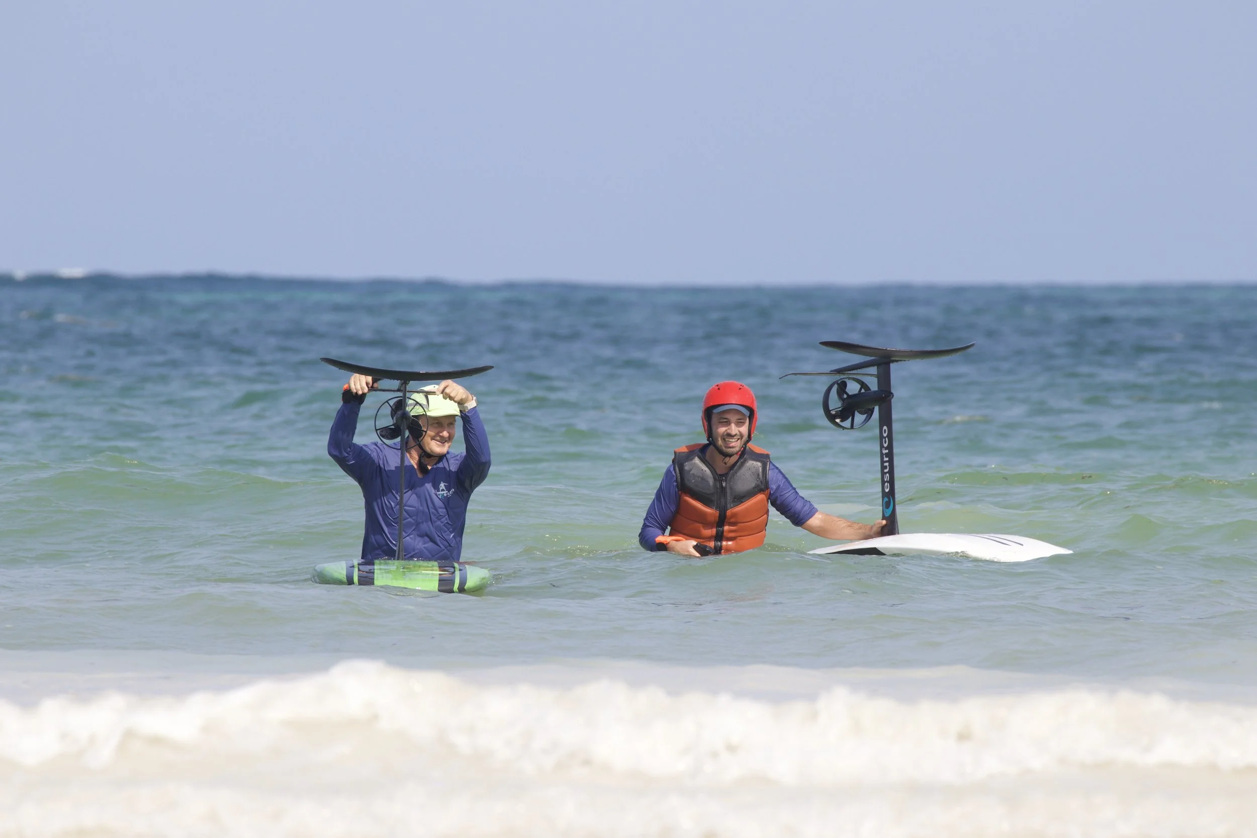 Two people in wetsuits, life jackets, and helmets, standing in the ocean holding surfboards with motorized sails, smiling and ready for surfing.