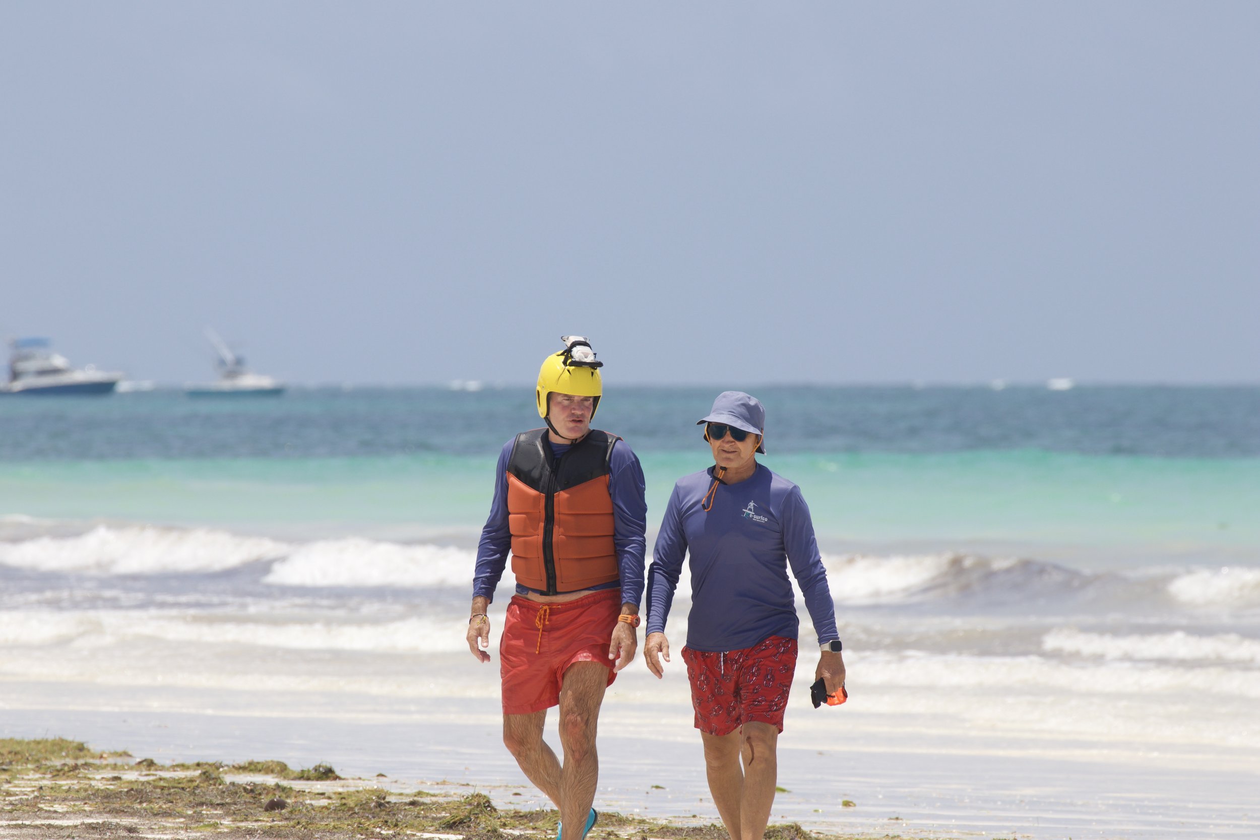 Two people walking along the beach, one wearing a yellow helmet and an orange life jacket, the other wearing a blue sun hat and sunglasses. The ocean and boats are visible in the background.