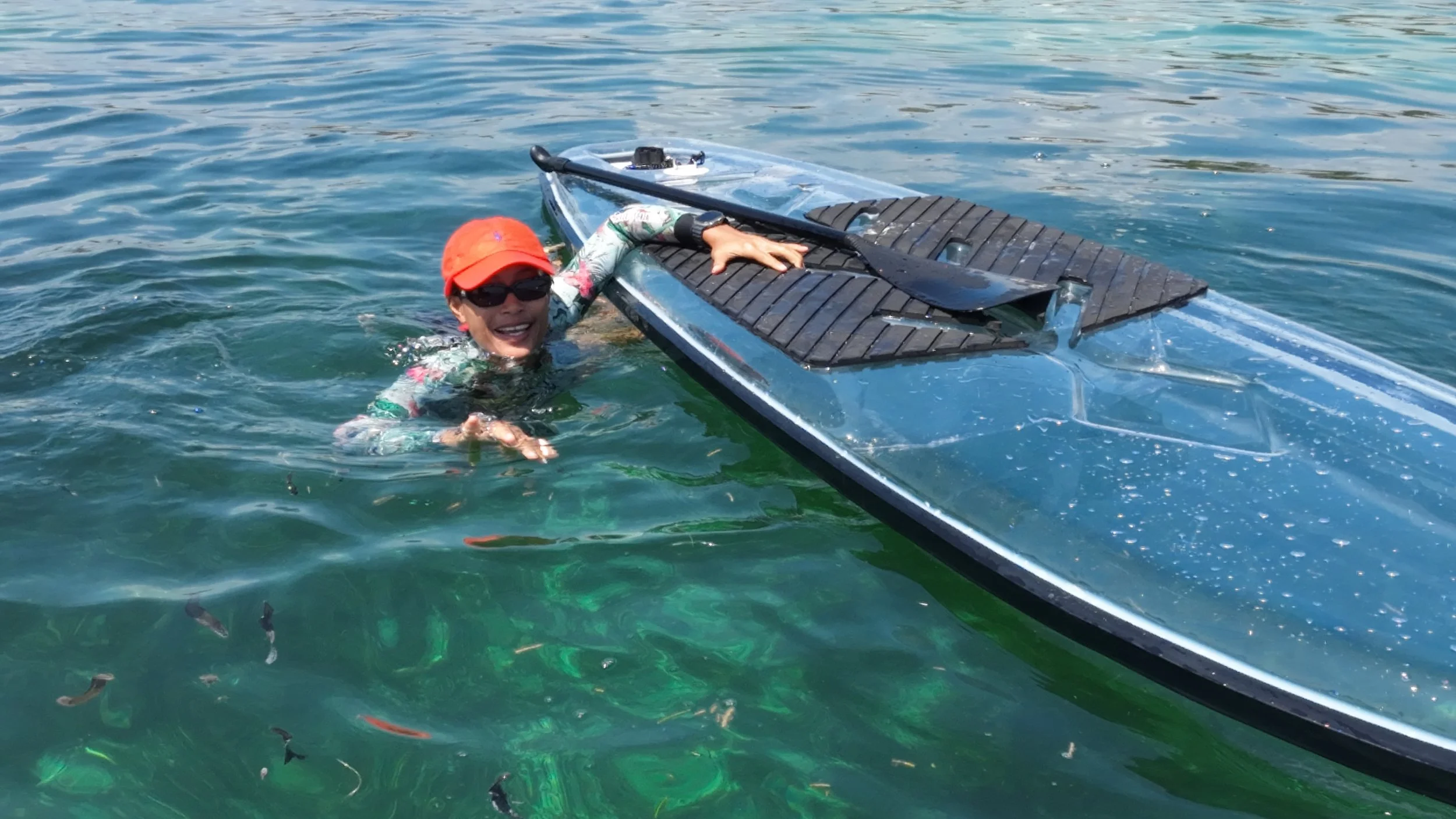 A woman with a red cap and sunglasses smiling in the water next to a black and silver kayak with a paddle resting on top.