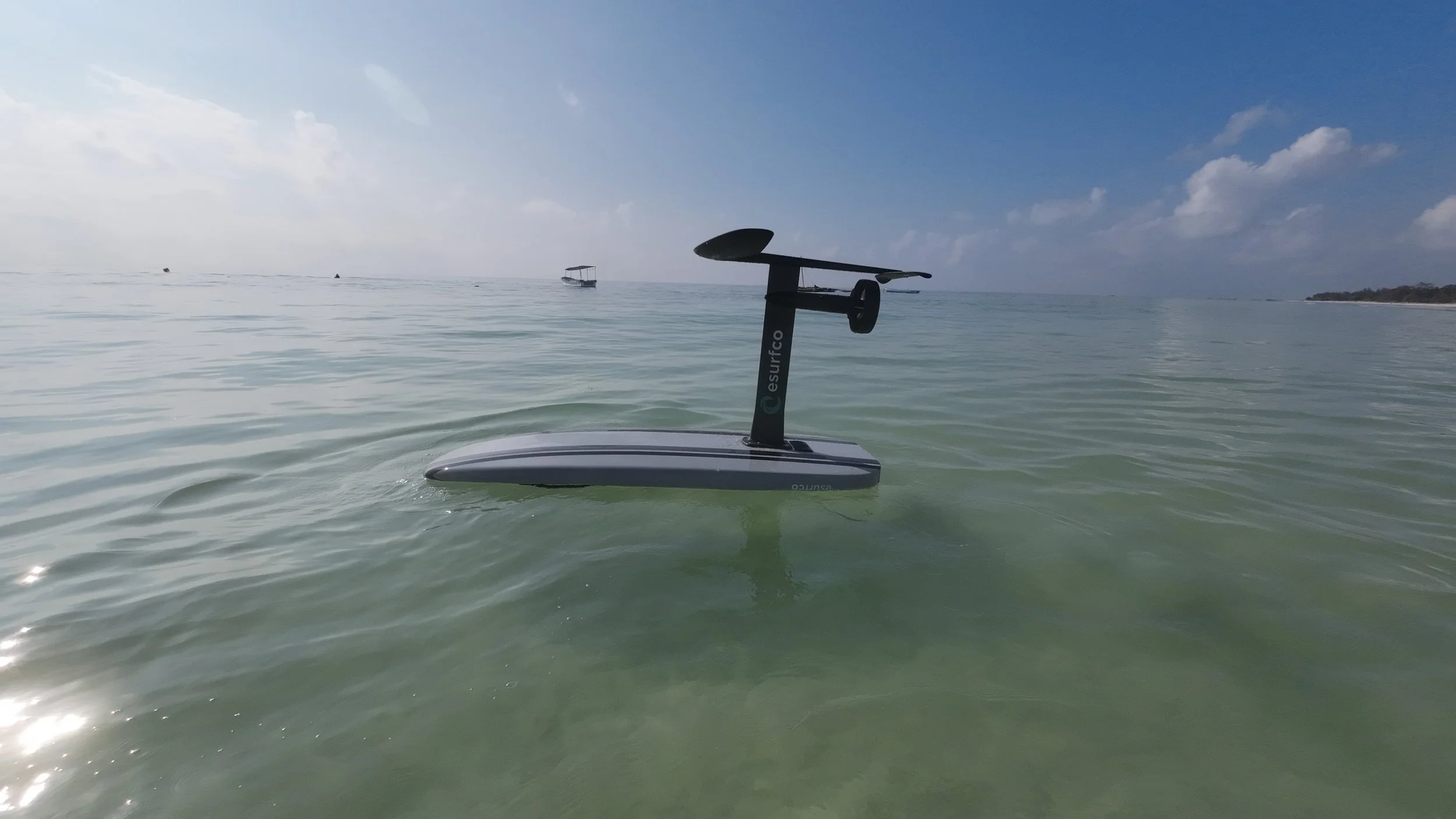 An electric surfboard with a detachable paddle floats on calm ocean water near the shore, with boats and a distant coastline visible under a partly cloudy sky.
