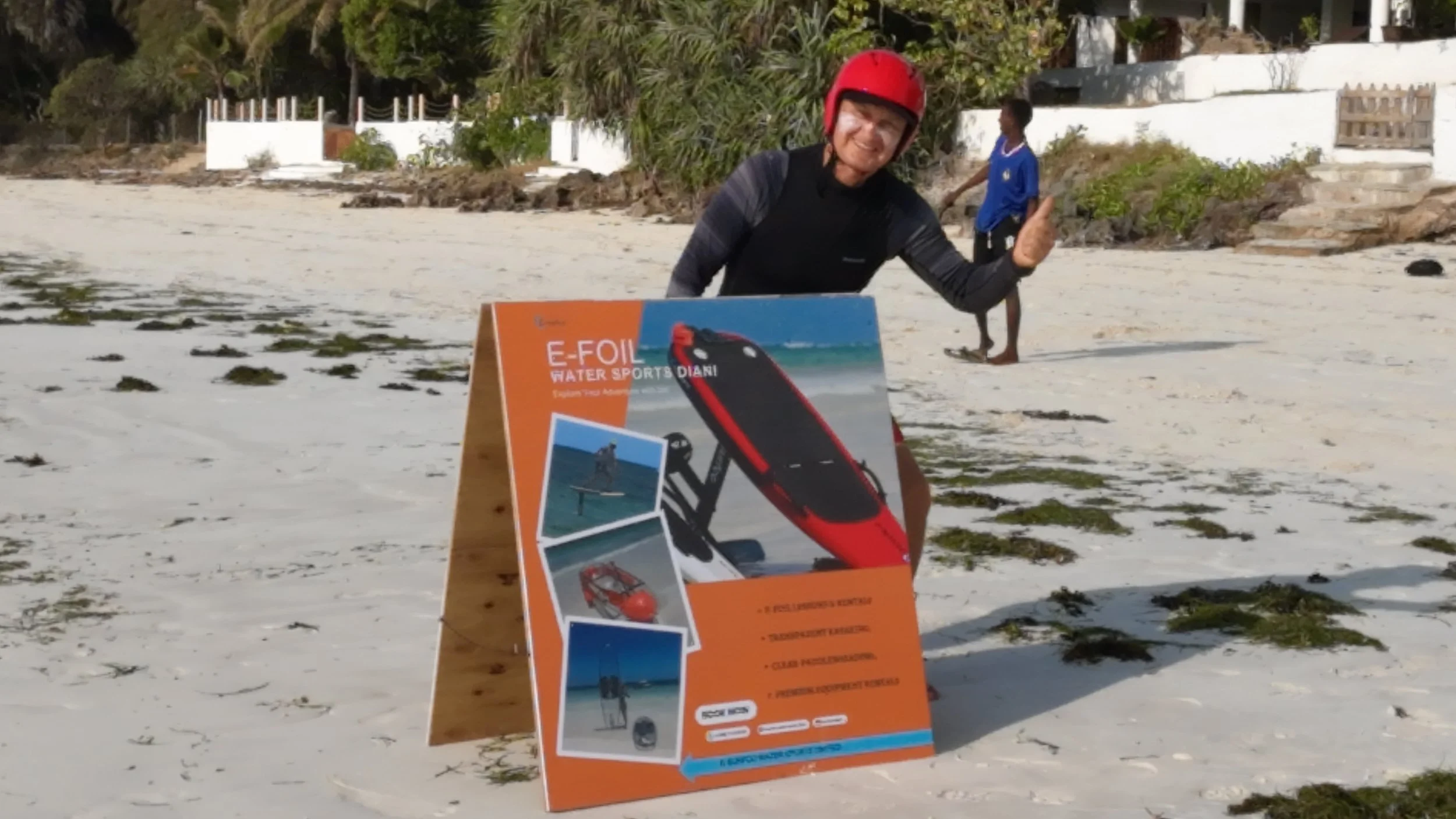 Man on the beach wearing a red helmet, giving a thumbs-up, standing next to an orange promotional sign for E-Foil water sports, with photo images of electric hydrofoil boards.