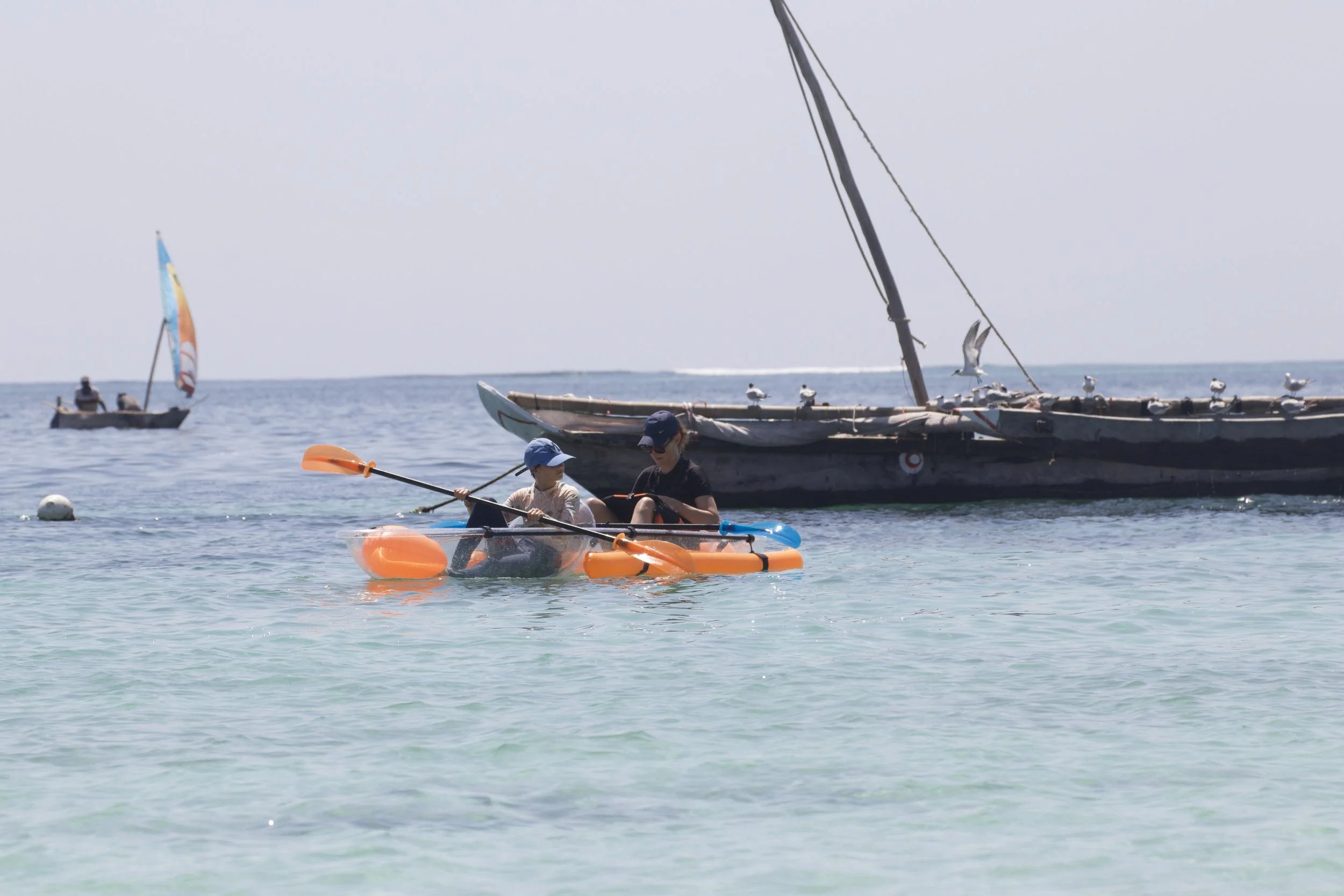 Two children paddling a small orange and gray kayak on clear, calm water with sailboats and seagulls in the background.