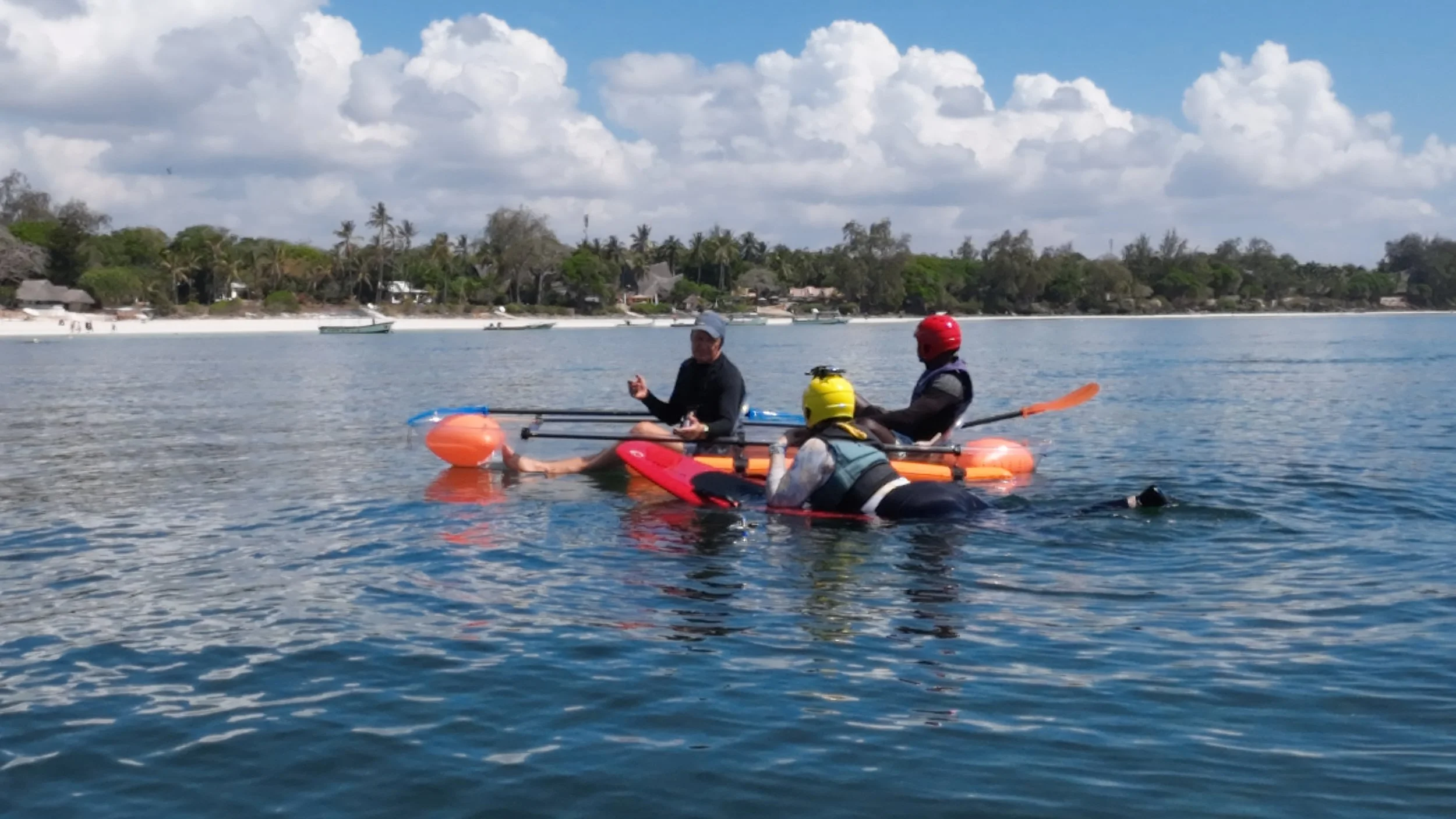 Three people on a kayak in a body of water, with a coastal shoreline and houses in the background. One person is in the water holding on to the kayak, two others are seated on the kayak, all wearing helmets and life vests, with the person on the righ