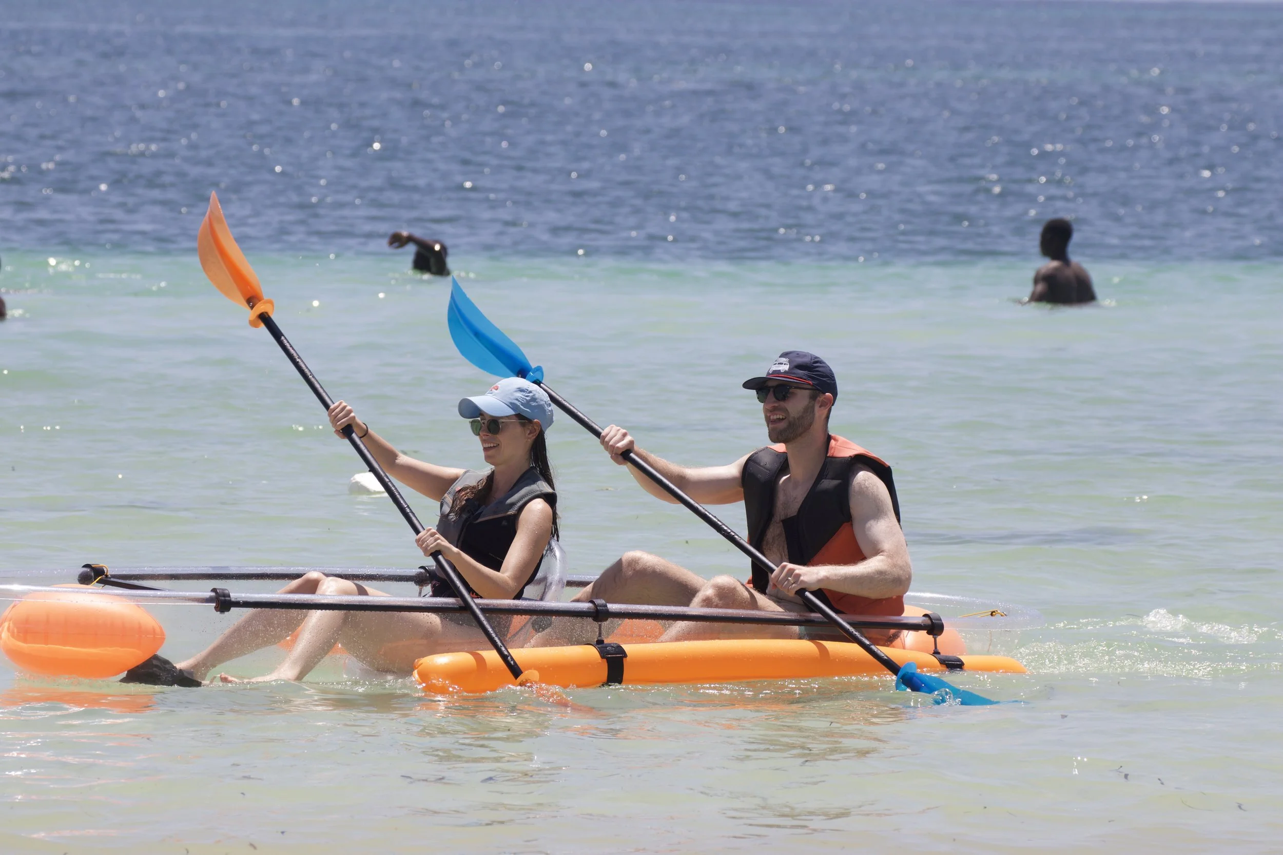 A smiling man and woman kayaking together on the ocean, both wearing sunglasses and life jackets, with others swimming in the background.