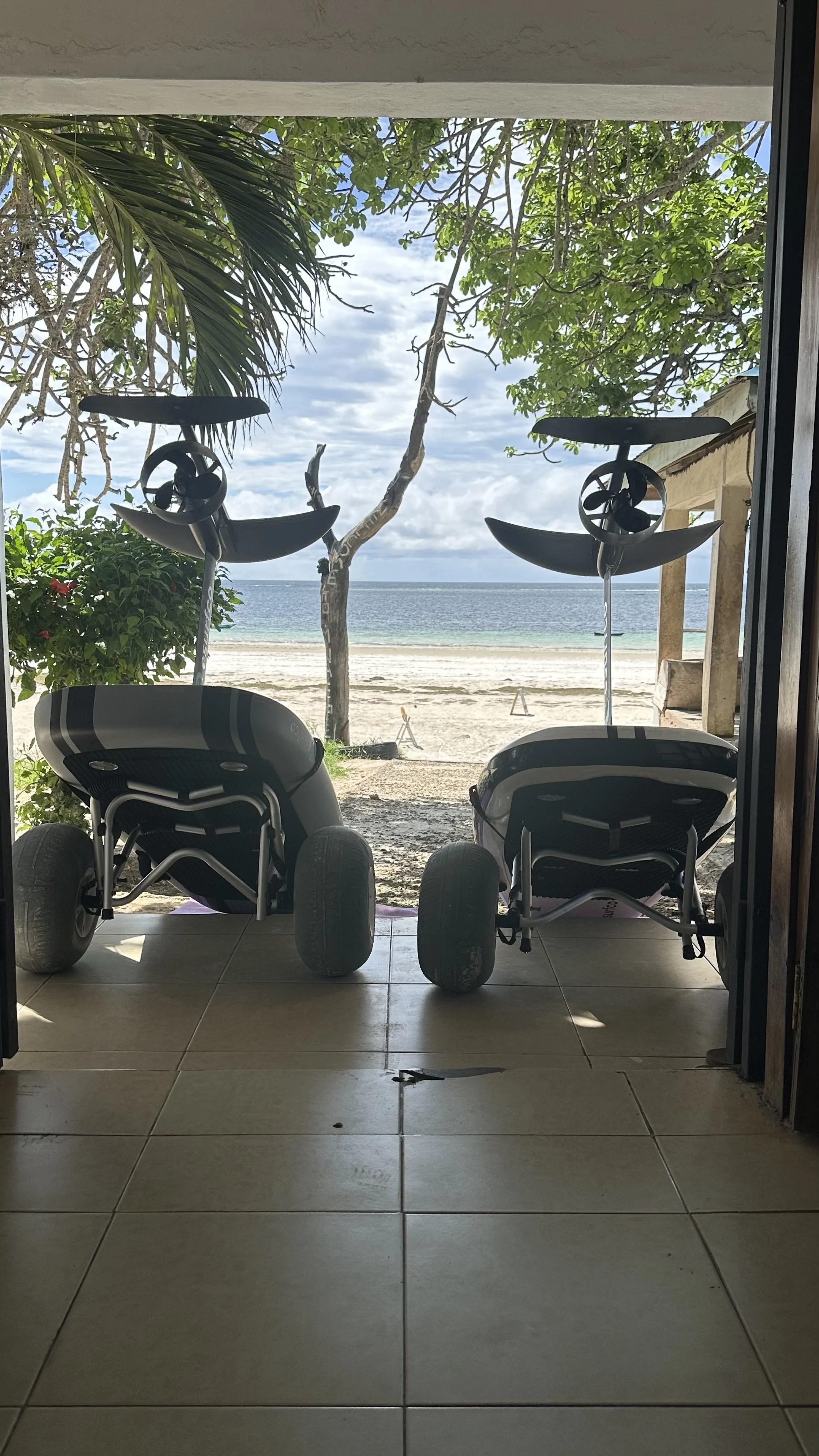 Two paddle boards leaning against a porch, facing the beach with trees and ocean in the background.