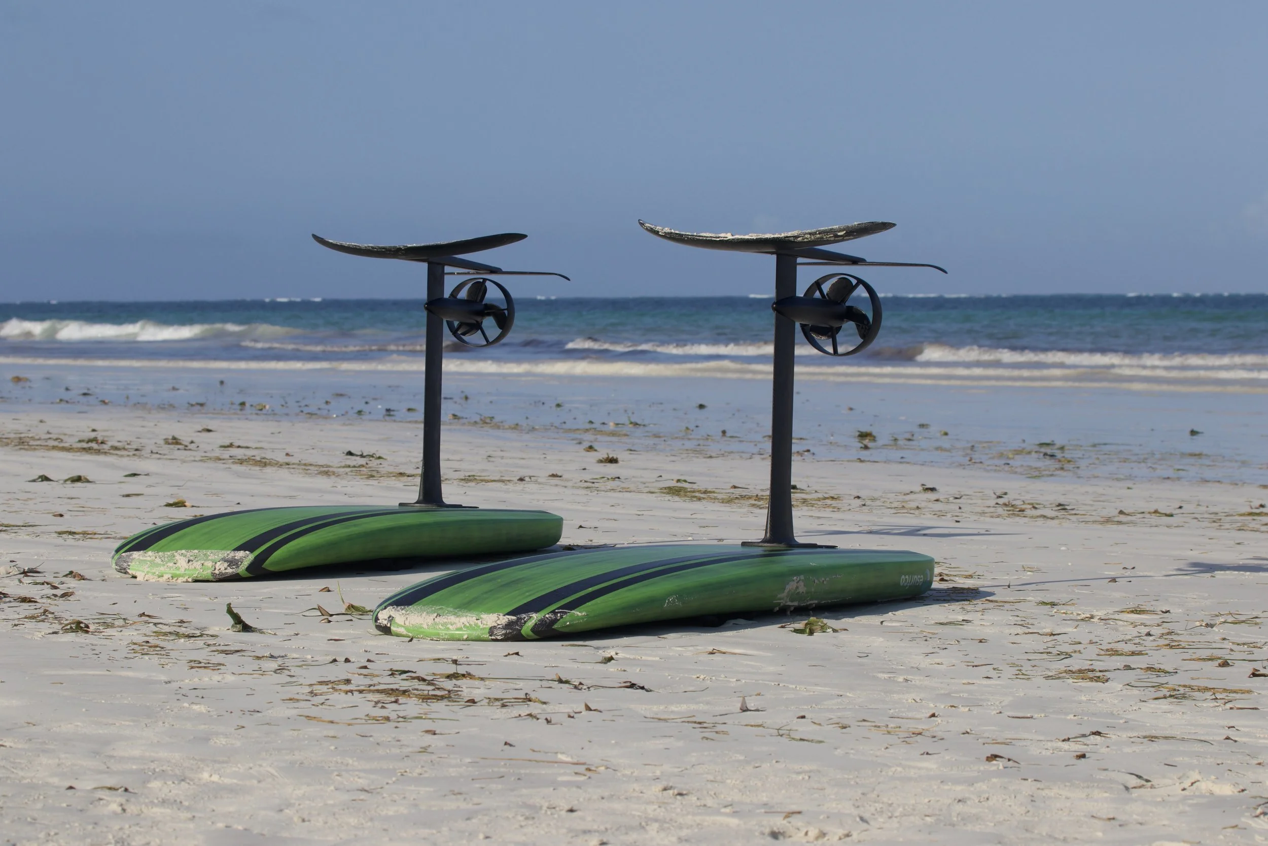 Two green paddleboards with black handles and black fins are lying on a sandy beach near the ocean. The sky is clear with some clouds and gentle waves are visible in the water.