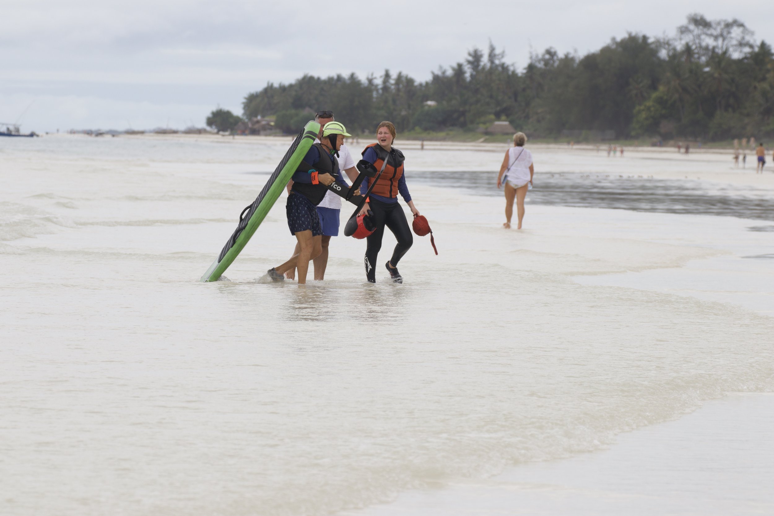 Four people at the beach, one carrying a paddleboard and two holding life jackets, walking along the shallow water near the shoreline, with another person in the background walking away.