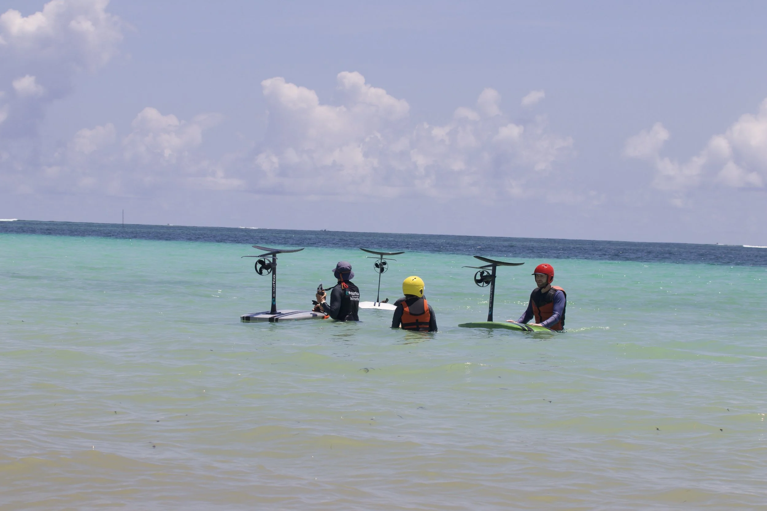 Three people in life jackets and helmets stand in shallow ocean water with windsurfing boards upright behind them, preparing for windsurfing activity, with a partly cloudy sky overhead.