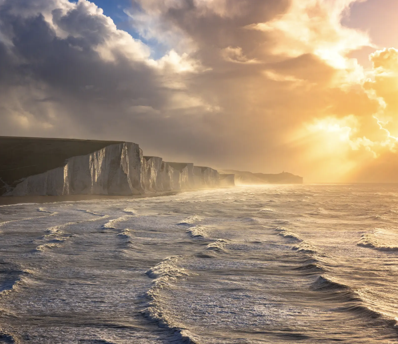 Cliffs along the coastline with wave-tossed water and a dramatic sunset sky with clouds and sunlight breaking through.