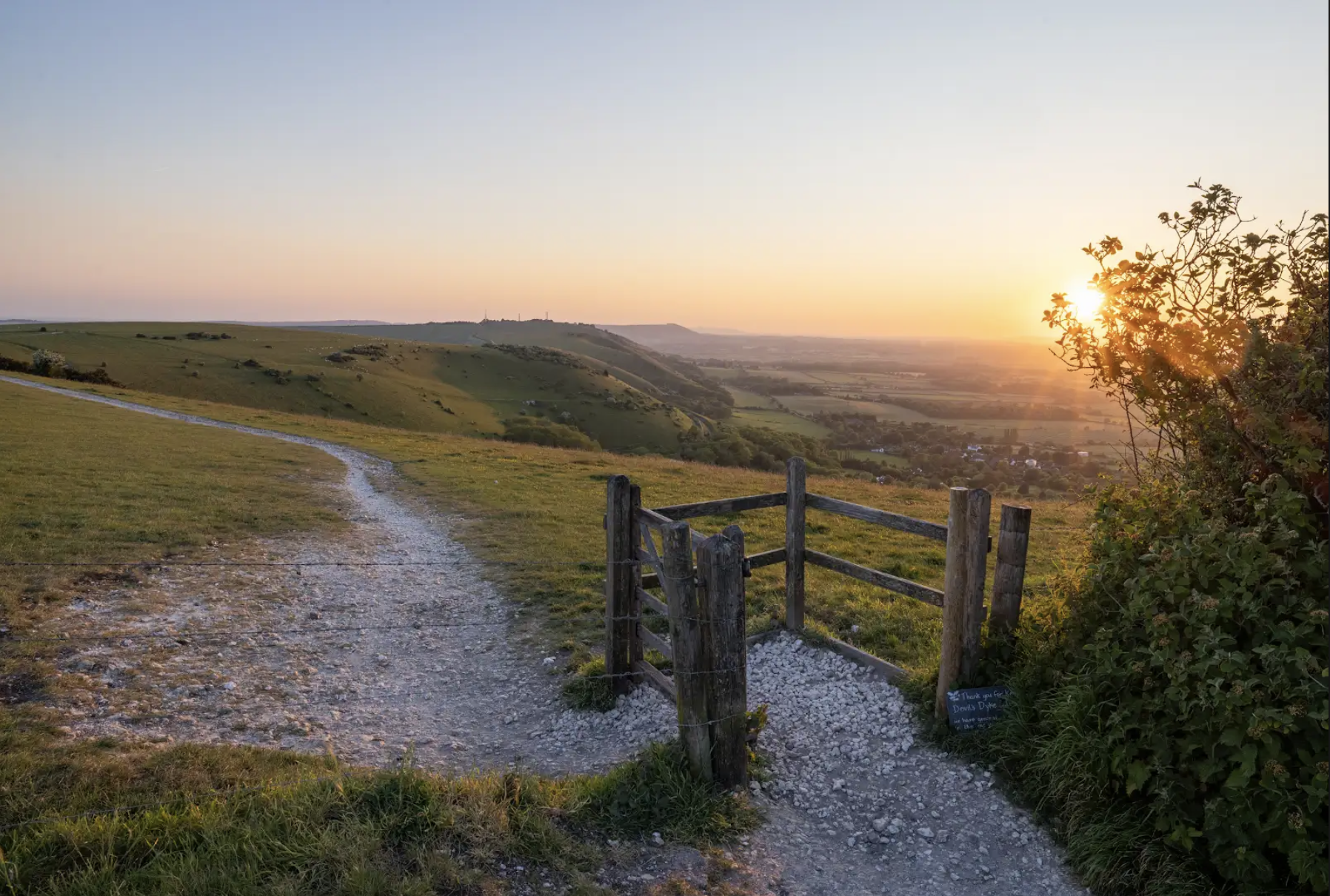 Sunset over rolling green hills with a dirt path and wooden fence in the foreground, a tree on the right with sunlight filtering through its leaves.
