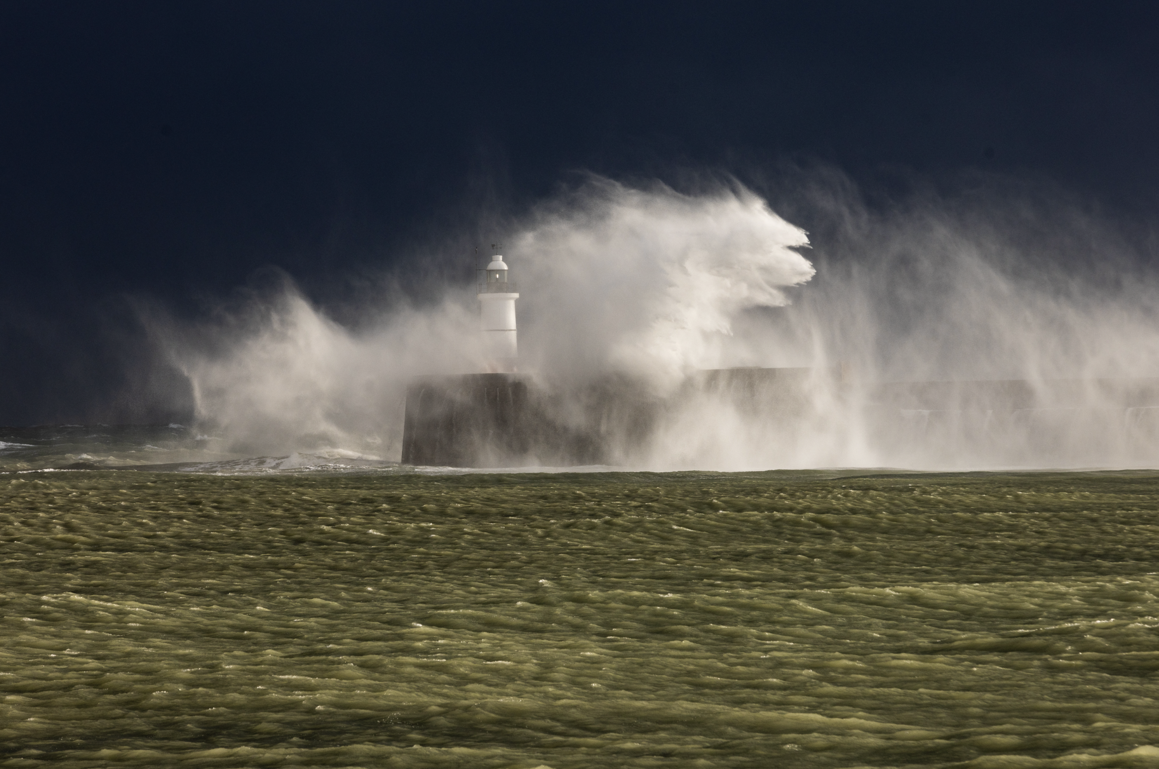 A lighthouse on a rocky coast with waves crashing against it, sending water and spray into the air during a stormy night.