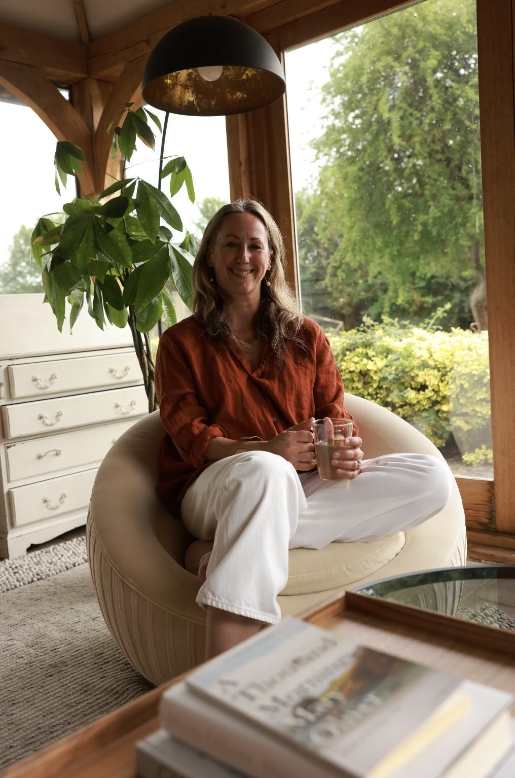 Leona Otty sitting in a cozy living room by a large window with green foliage outside, holding a glass mug, smiling at the camera, with a large potted plant behind her and a white dresser to the side.