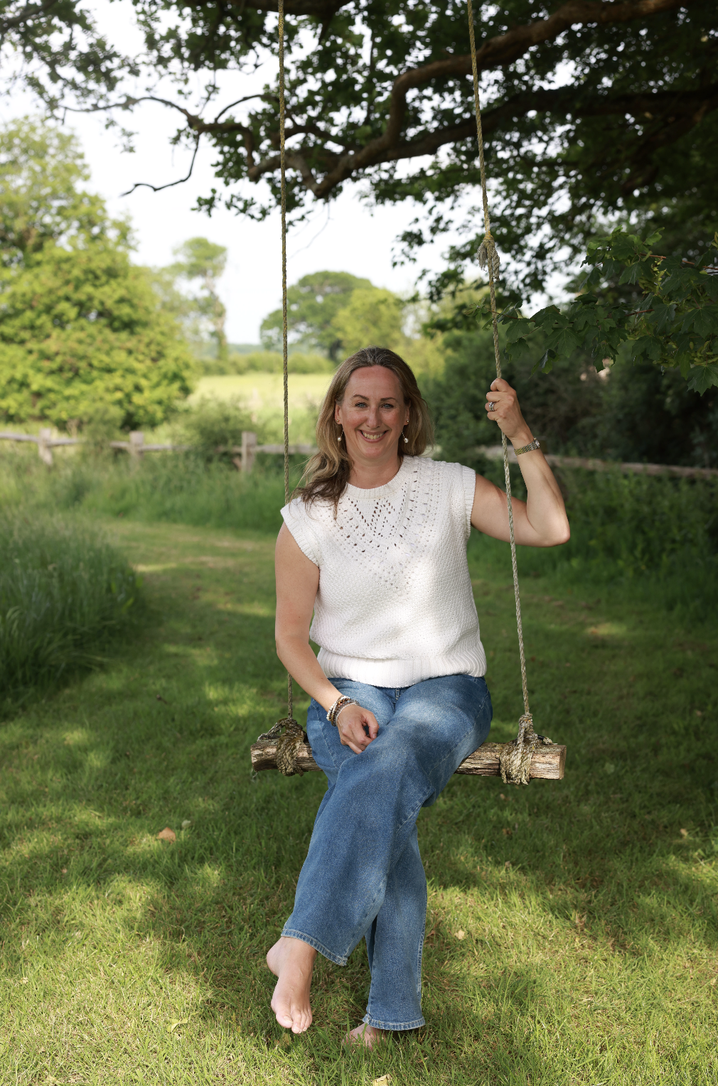 Leona Otty sitting on a wooden swing hanging from a tree branch in a lush green outdoor setting, smiling at the camera.