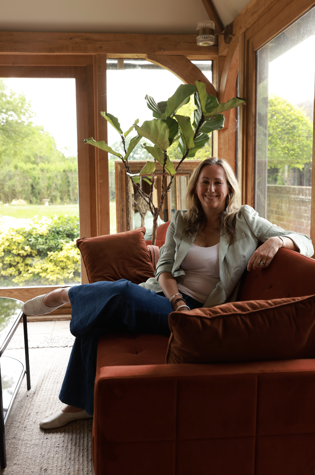 Leona Otty with blonde hair, wearing a white top and beige blazer, sitting on a rust-colored sofa in a room with large windows, natural light, and greenery outside. Behind her is a tall potted plant.