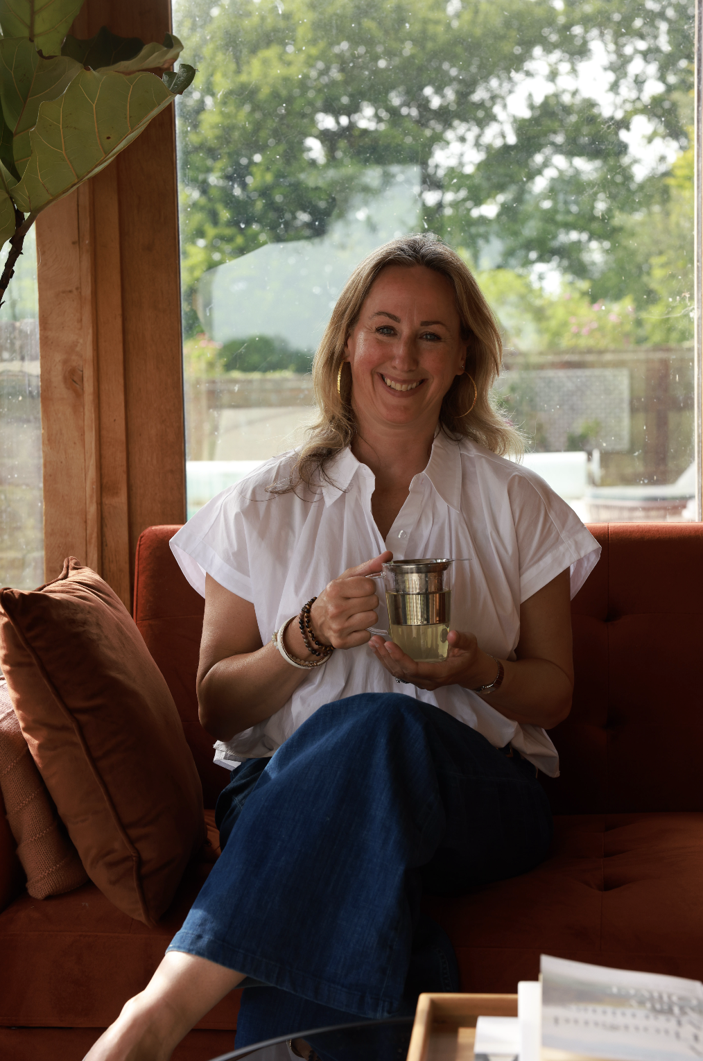 Leona Otty sitting on a burnt orange couch holding a clear mug with tea, smiling at the camera, with a large window and green trees in the background.