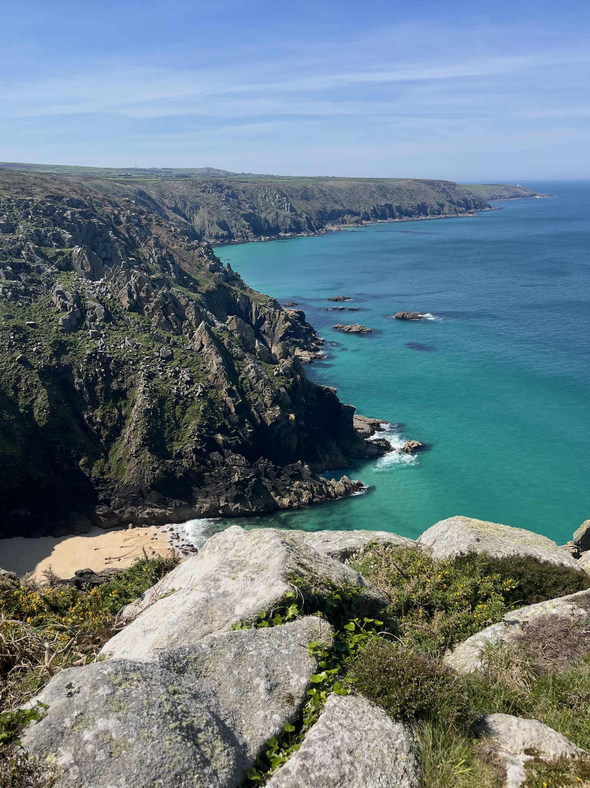 Coastal cliffs overlooking a turquoise bay with a small sandy beach, rocky shoreline, and green vegetation under a blue sky.