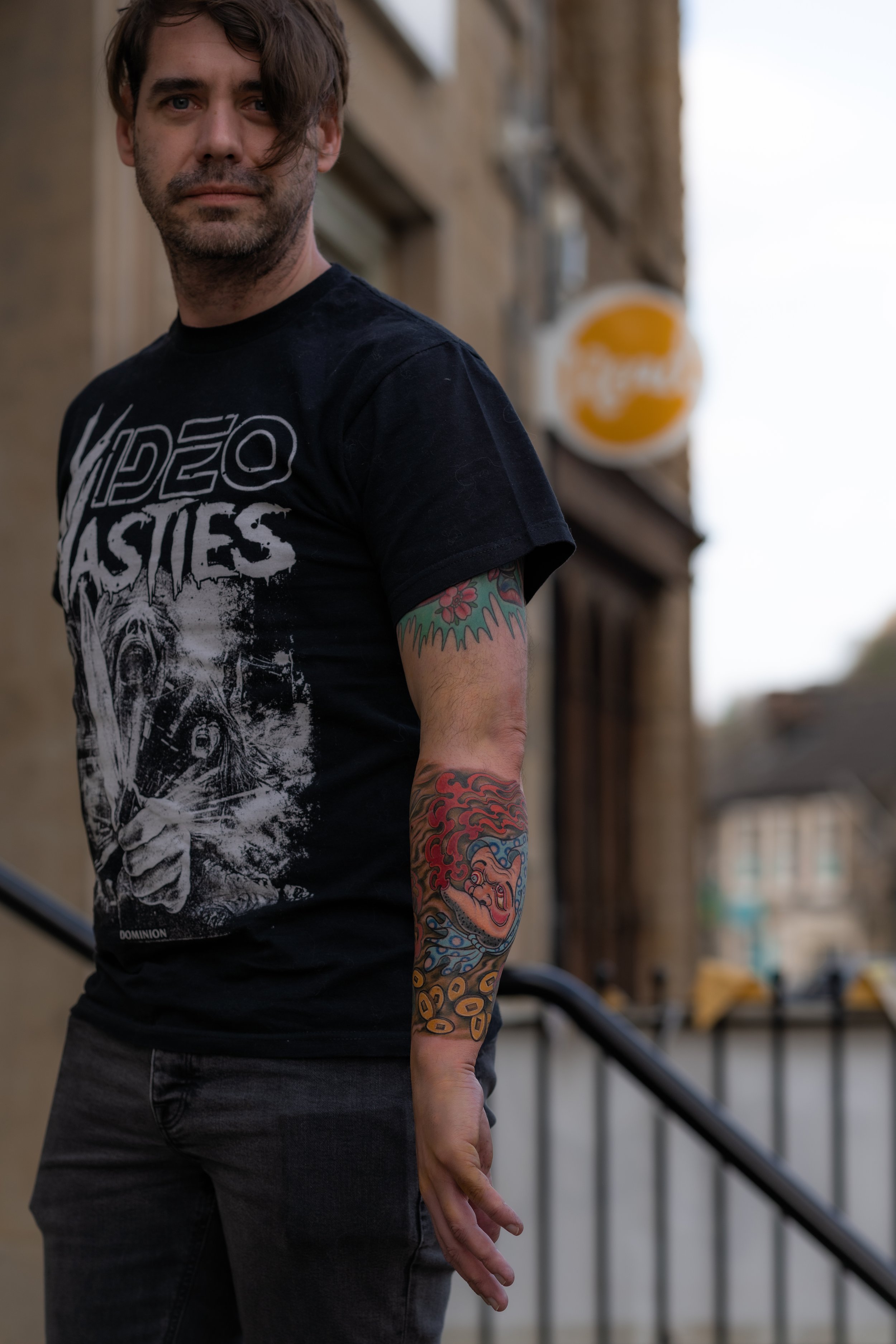 A man with dark hair and tattoos on his right arm stands outdoors, wearing a black graphic T-shirt with the phrase 'Video Bashes' and a black pair of pants. He is looking towards the camera with a neutral expression, with a building and a sign in the background.