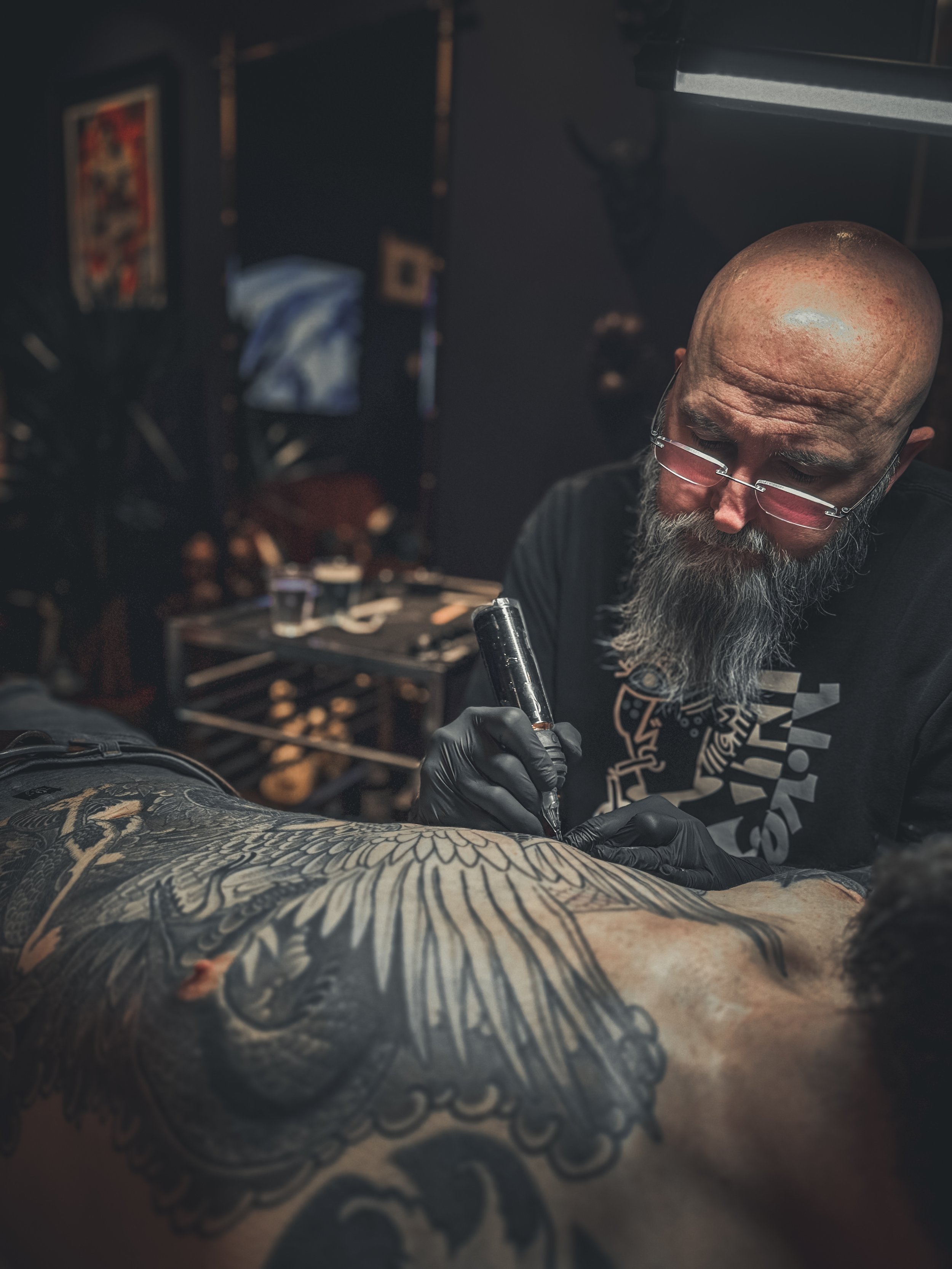 A tattoo artist with a bald head, glasses, and a beard tattooing a large design on a person's back in a dimly lit studio.