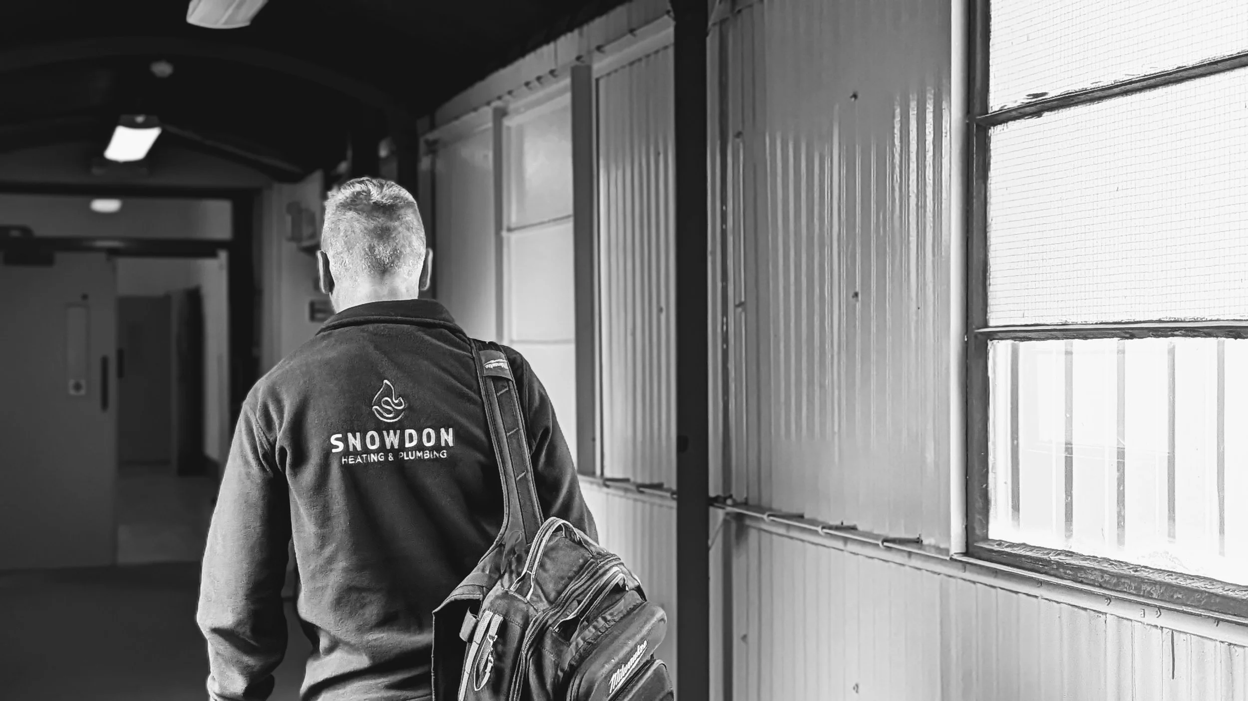A worker seen from the back, wearing a jacket with the logo 'Snowdon Heating & Plumbing' and carrying a backpack, walking inside an industrial or warehouse building with large windows and metal walls.