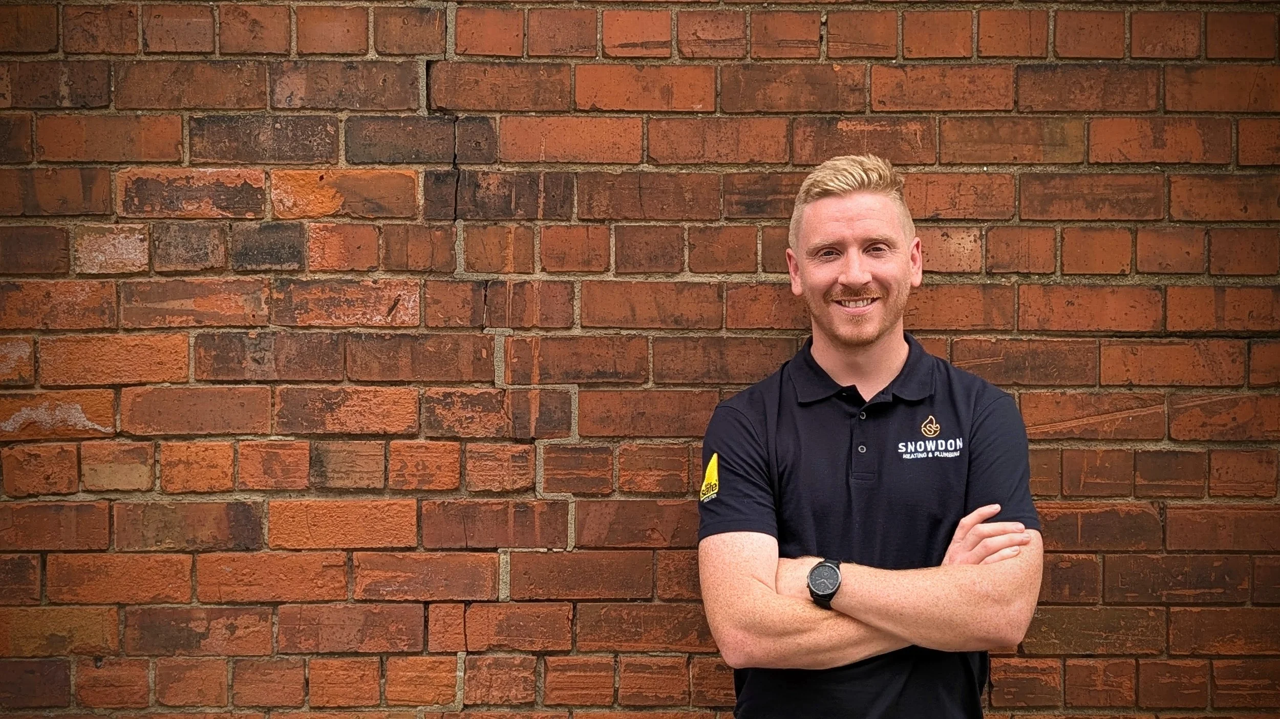 A man with short blond hair and a beard is standing with his arms crossed against a red brick wall. He is wearing a black polo shirt with logos, one reading 'SNOWDON Heating & Plumbing' and the other a yellow patch, possibly indicating safety or compliance.