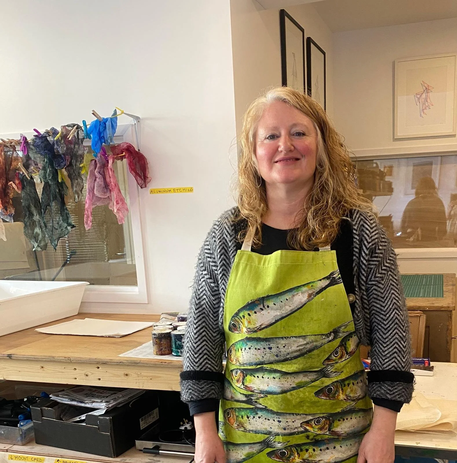 A woman with curly blonde hair wearing a fish patterned apron and a black and white cardigan, standing in a workshop or studio. Behind her is a workbench with jars of paint and other art supplies, and a display of colorful fabric or paper scraps hanging on a line. There are framed pictures on the wall and a window with reflections.