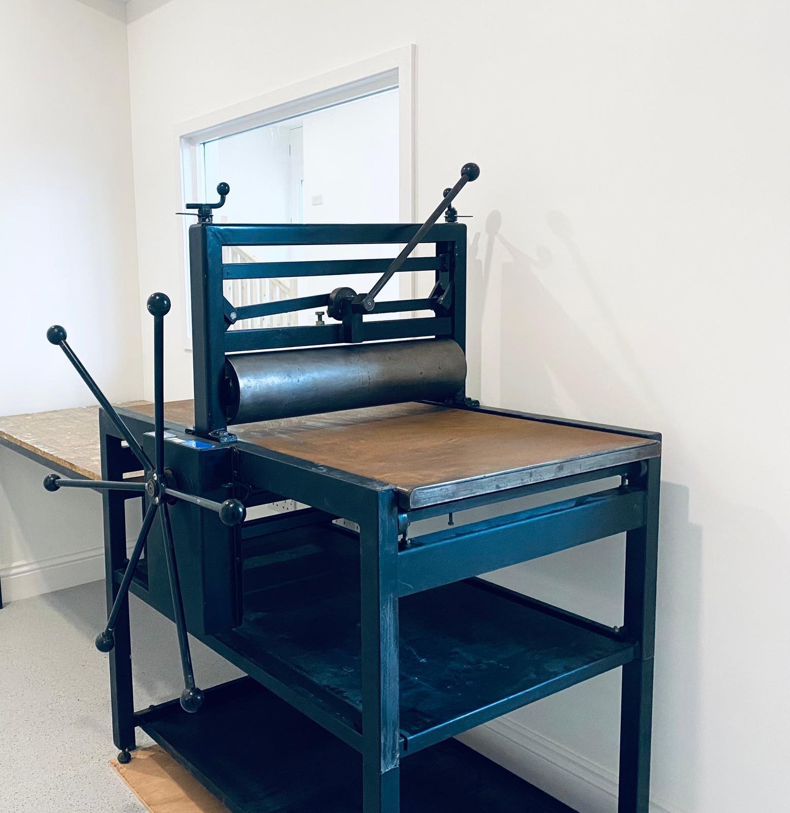 A vintage printing press with a wooden surface, black metal frame, and various levers and parts, situated in a room with white walls and a window.