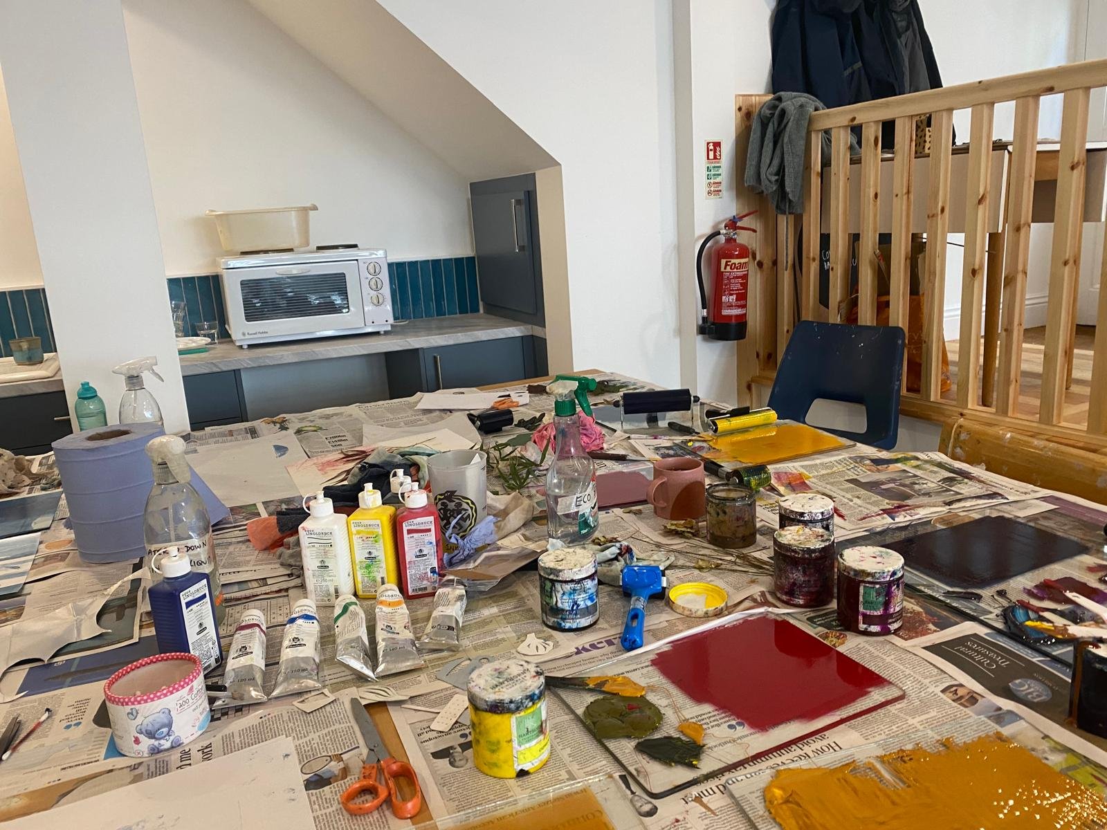 A cluttered table covered with newspapers, paint supplies, and art tools, with a water spray bottle, paint tubes, jars, scissors, and a red notebook, in a room with a blue kitchen area and a wooden staircase railing.