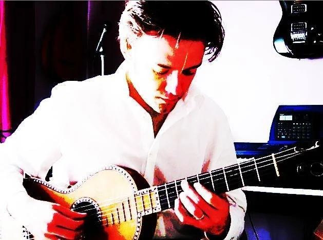 Man with dark hair playing an acoustic guitar in a room.