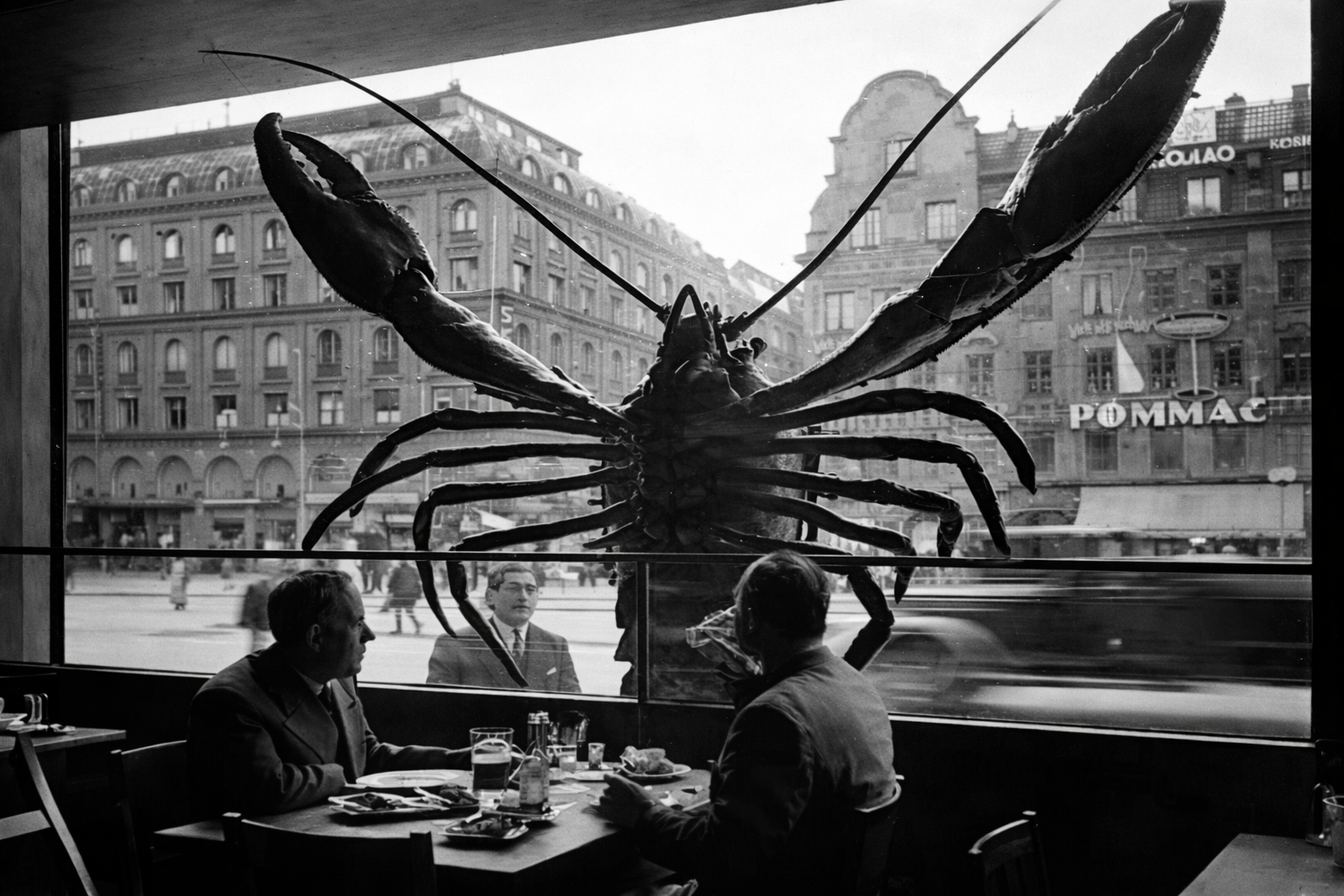 A giant lobster sculpture sitting on the other side of a restaurant window with two men dining inside. Outside city buildings and pedestrians are visible.