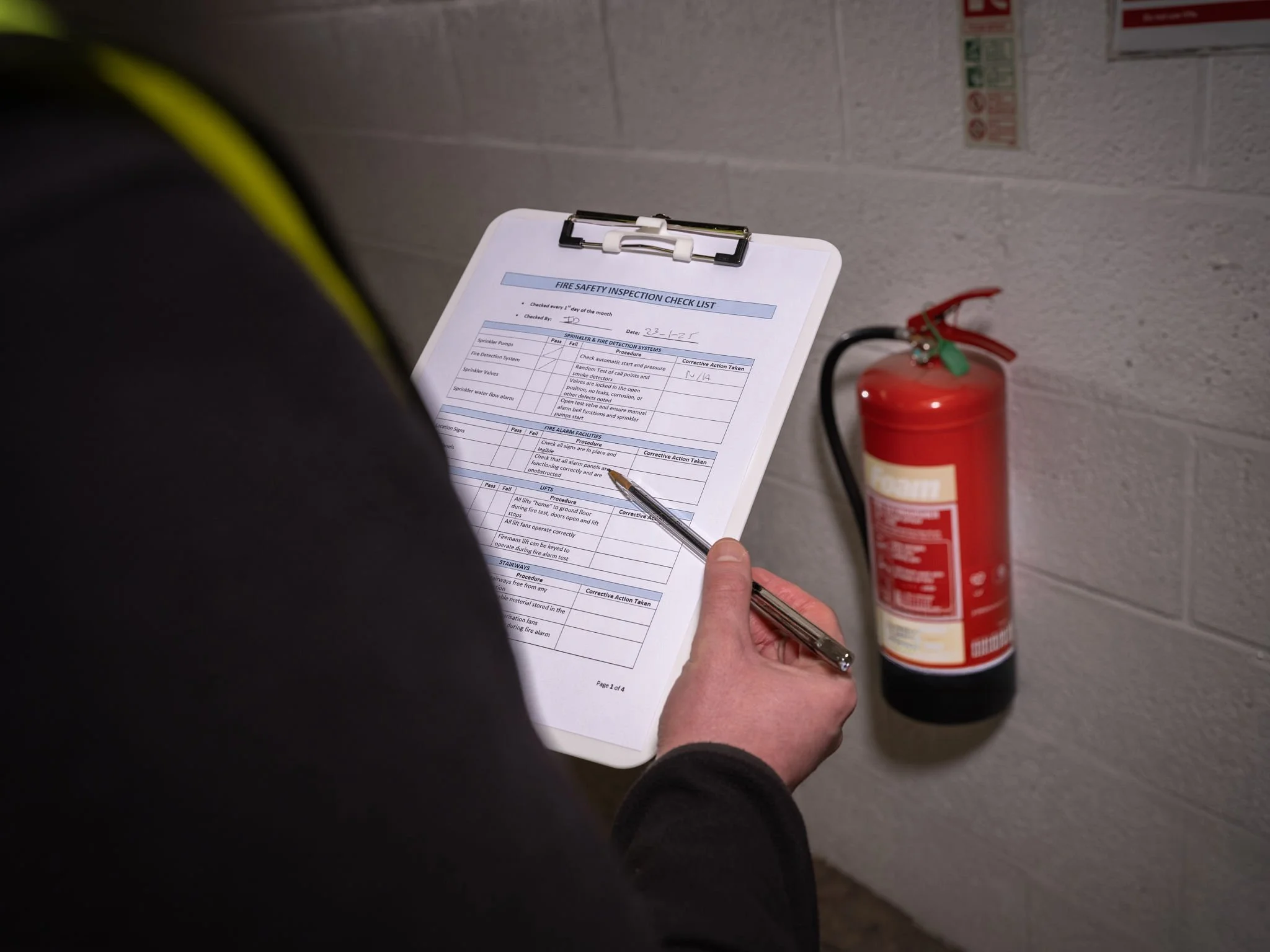 A person holding a checklist on a clipboard and a pen, inspecting a fire safety checklist near a red fire extinguisher mounted on a gray brick wall.
