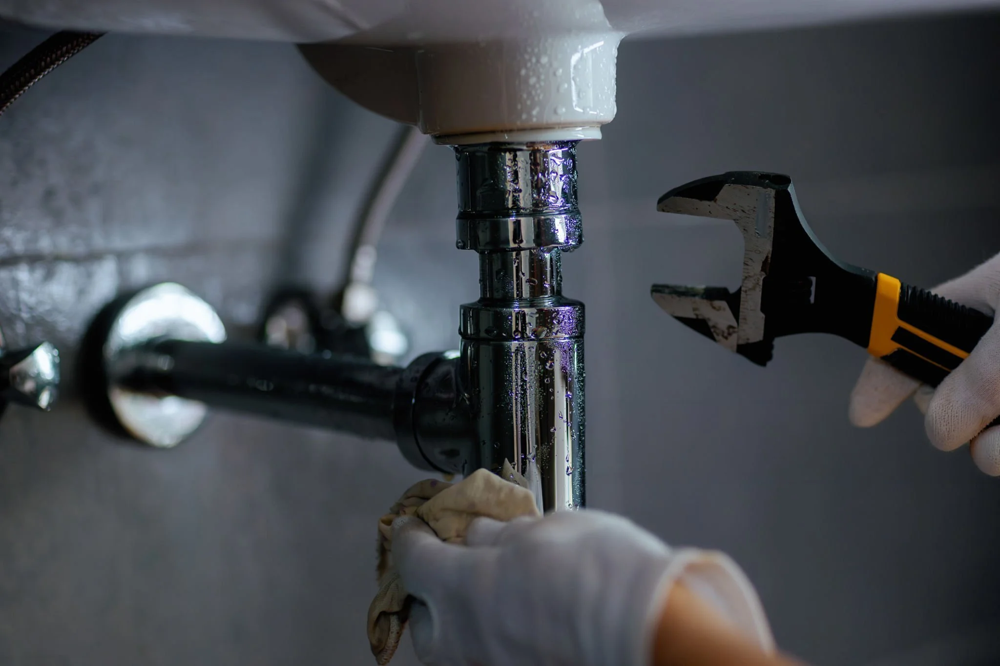 A person wearing gloves is using a wrench to tighten or loosen a pipe underneath a sink, with water droplets on the pipe.