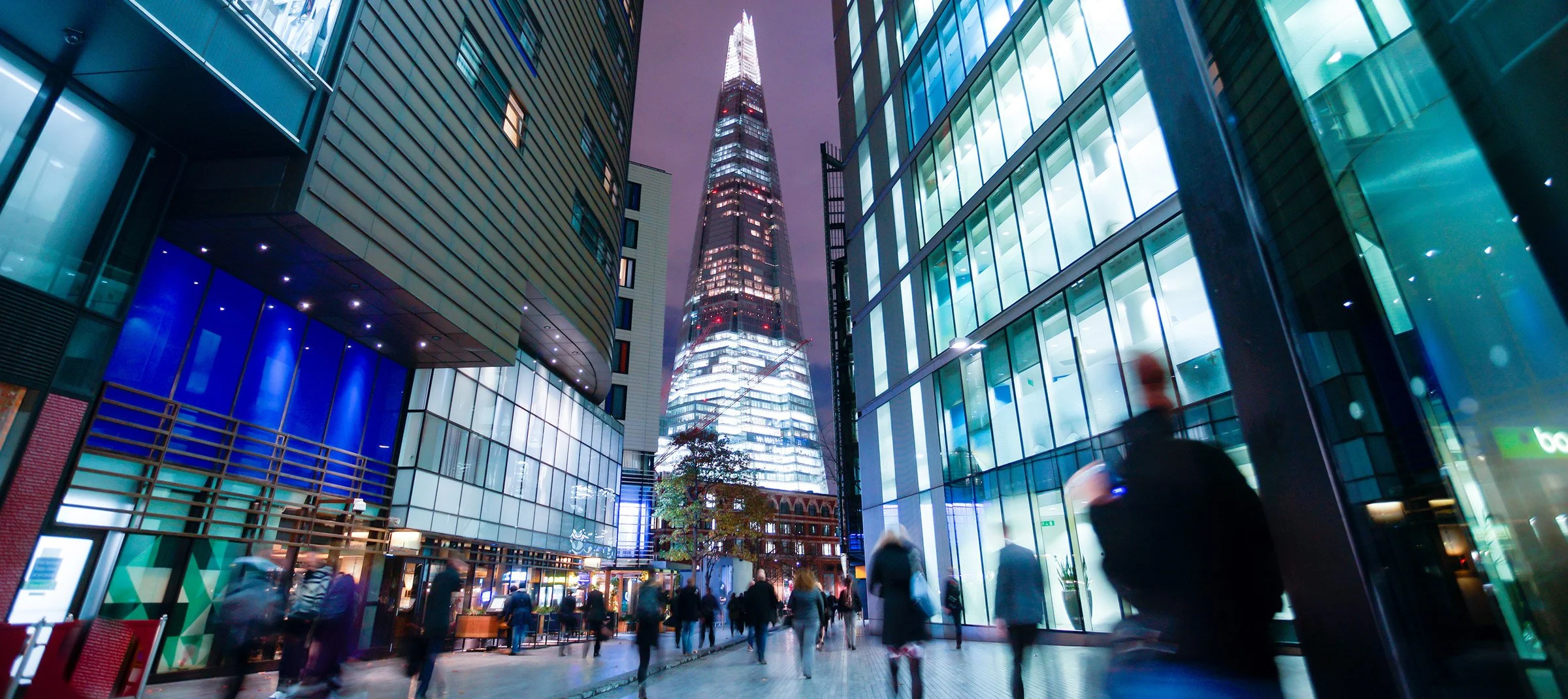 Night view of a city street with tall, illuminated skyscrapers, including the Shard building in London, reflective glass facades, and blurred pedestrians walking.