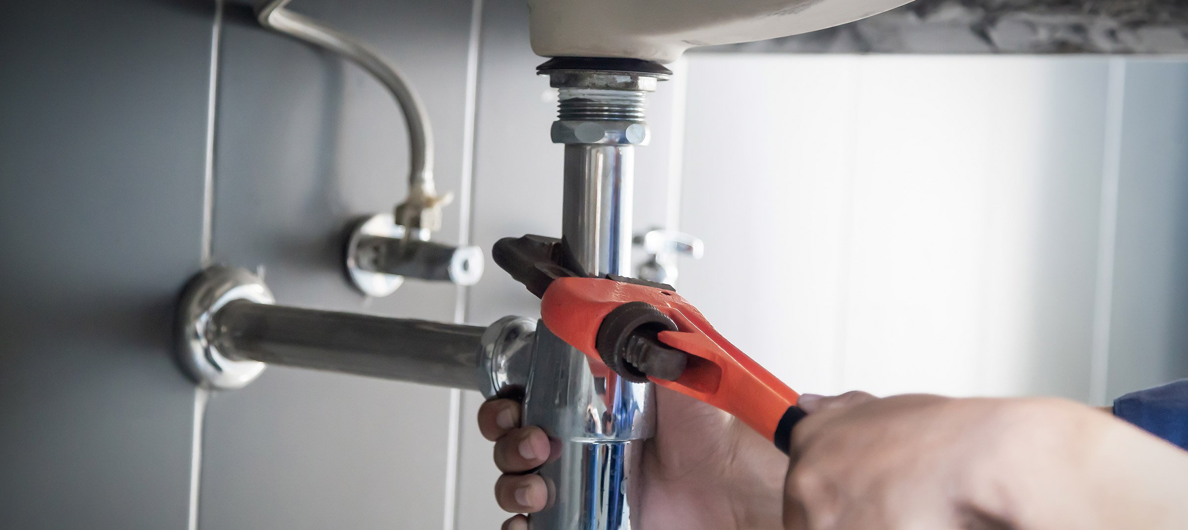 A person wearing gloves is using a wrench to tighten or loosen a pipe underneath a sink, with water droplets on the pipe.