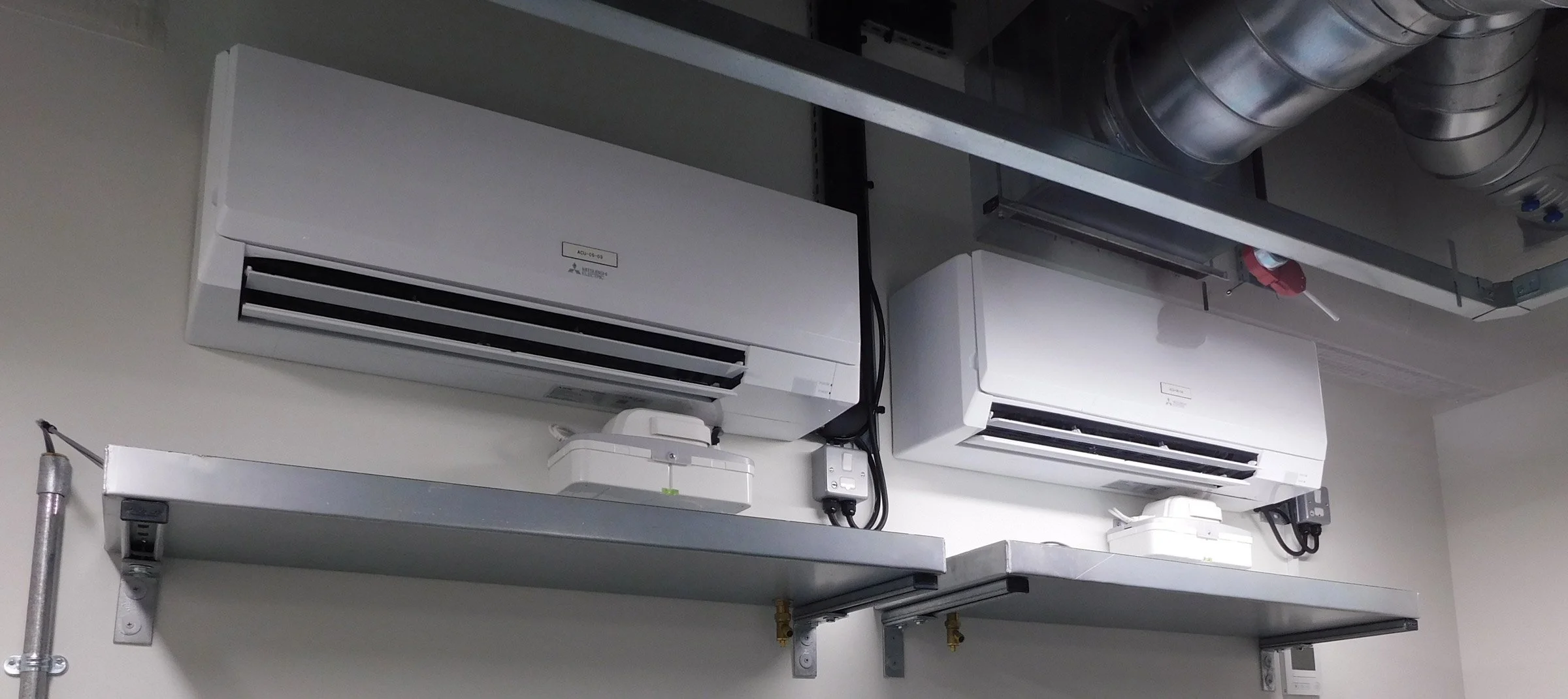 Two white wall-mounted air conditioning units below a metal duct system, with electrical wiring and a remote control on a white shelf.