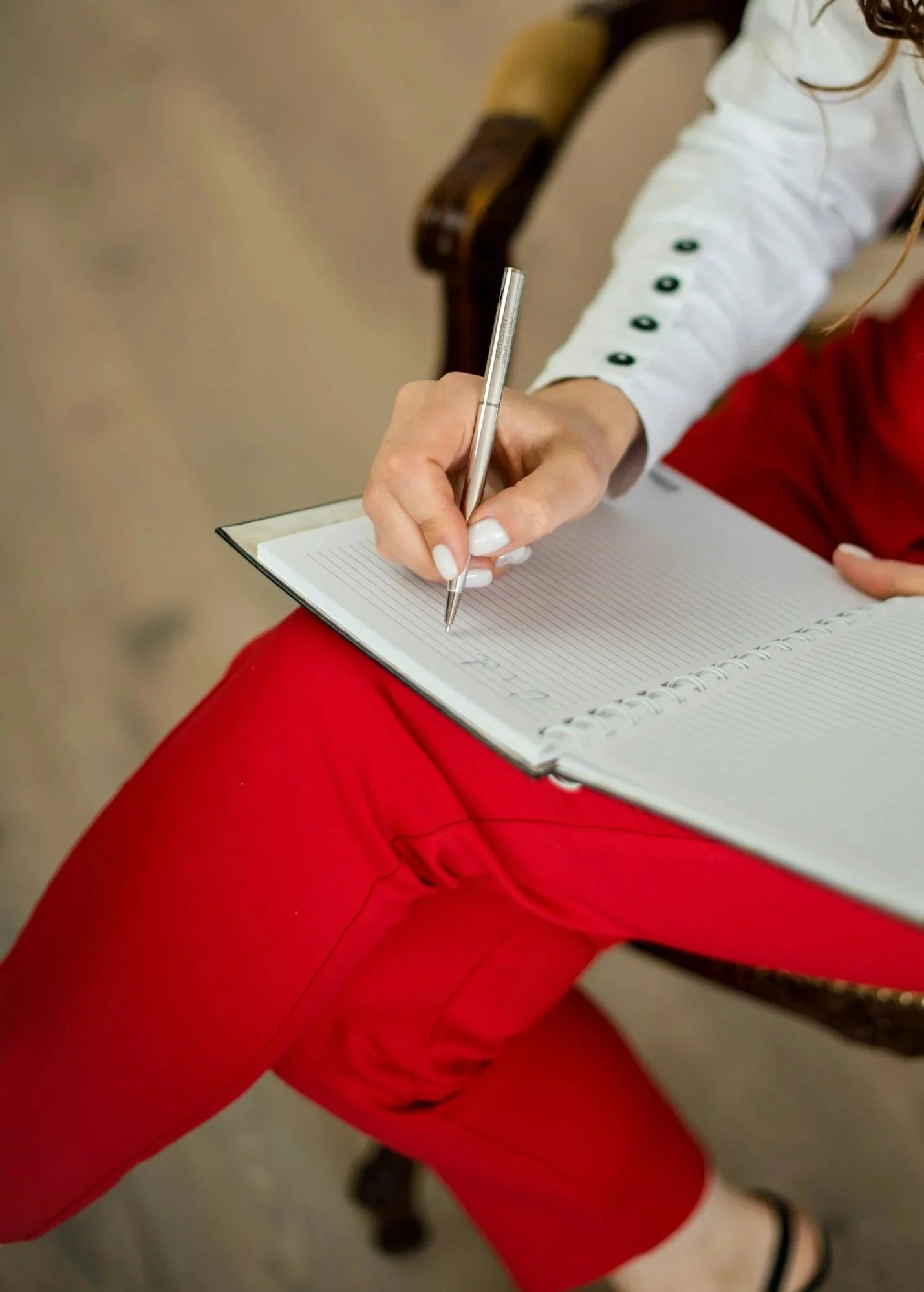 Eine Person sitzt auf einem Stuhl und schreibt in ein offenes Notizbuch mit einem silbernen Kugelschreiber. Die Person trägt eine weiße Bluse mit schwarzen Knöpfen und eine rote Hose.