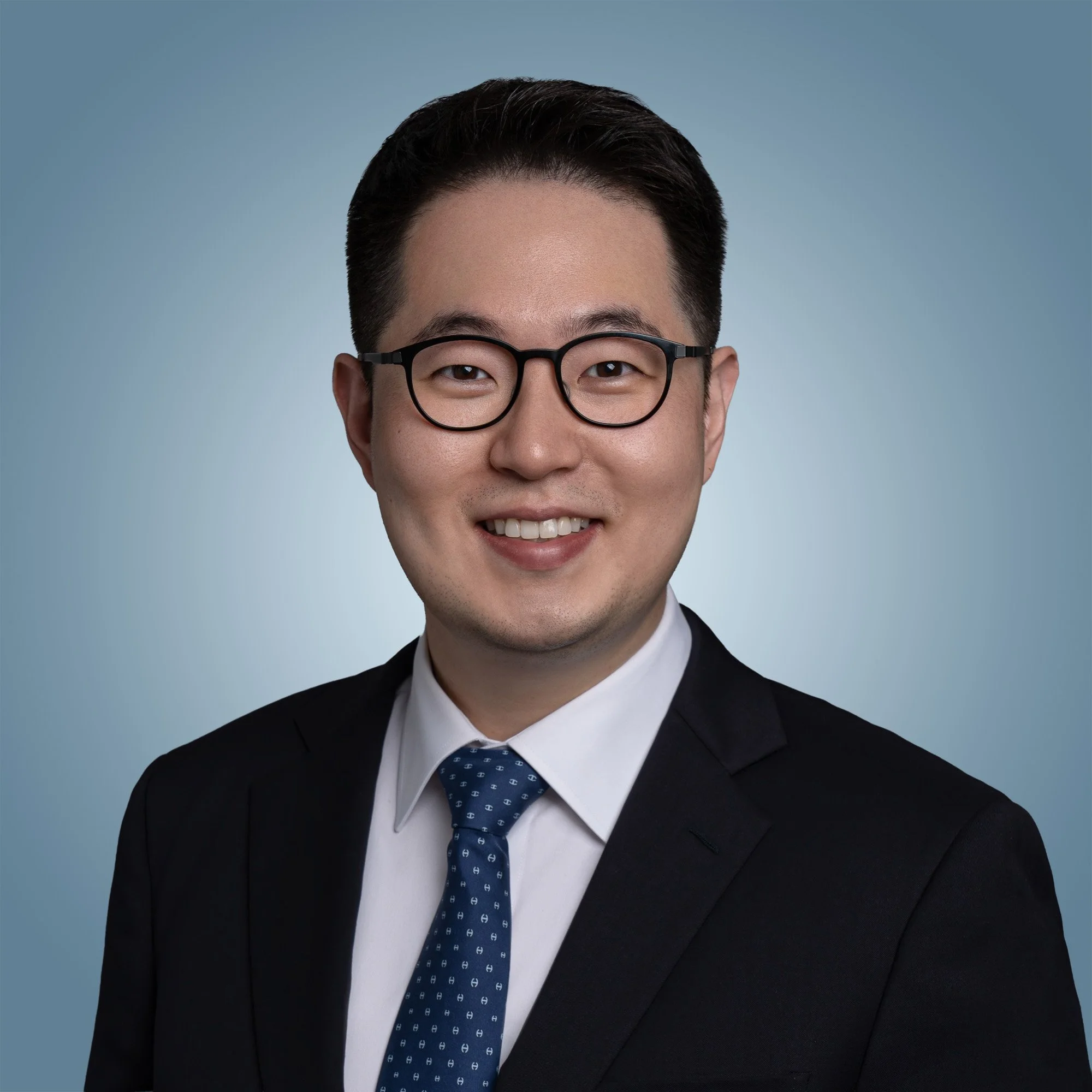 A man dressed in formal attire, wearing glasses, a dark suit, white shirt, and a blue tie with a pattern, smiling against a light blue background.