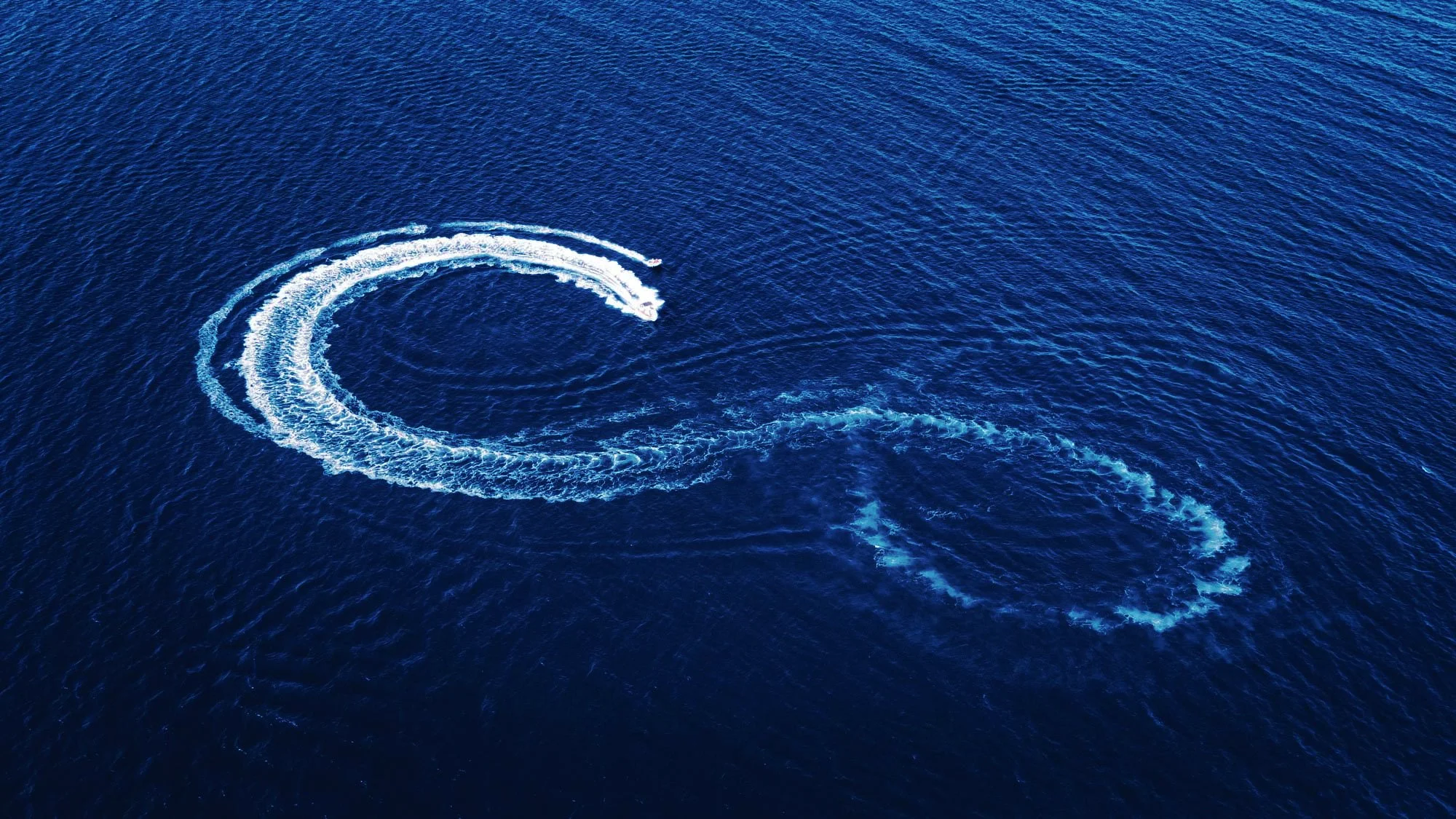 Two boats creating large spray and wake in the ocean, forming a curved pattern.