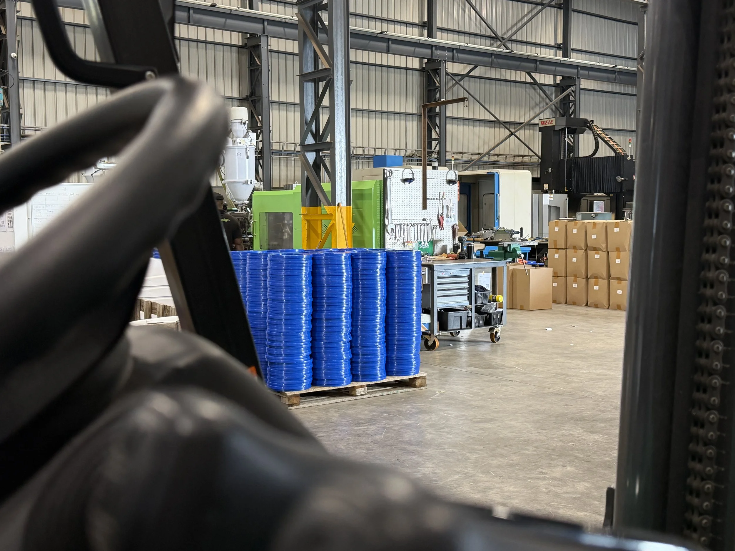 View of a warehouse interior with steel beams, shelves, and various equipment, partially seen through a chain-link fence or industrial pipes in the foreground.