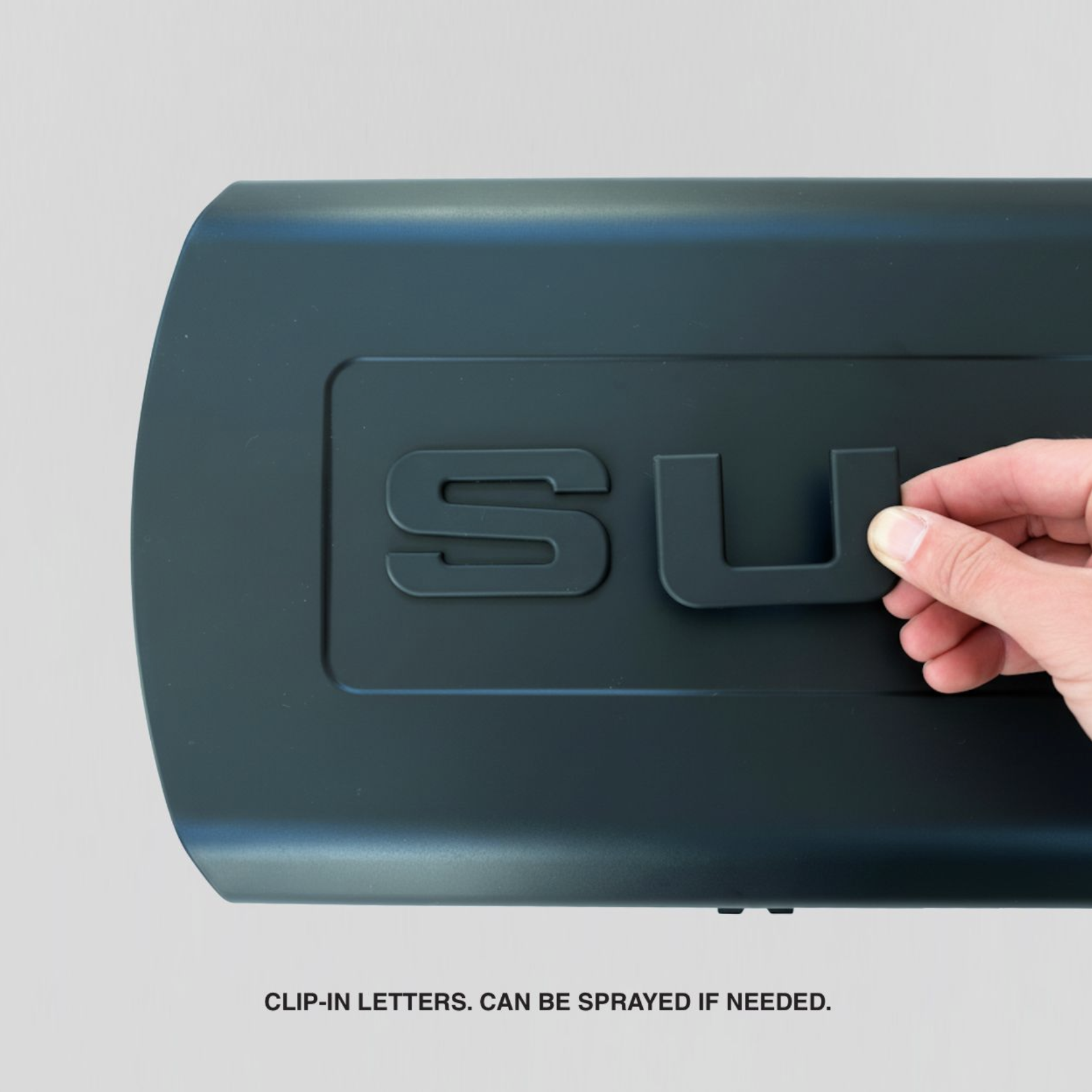 A close-up of a teal-colored electronic device with a hand holding a clip-in letter 'L' on its surface. The device has the letter 'S' already attached and text below states, 'CLIP-IN LETTERS CAN BE SPRAYED IF NEEDED.'
