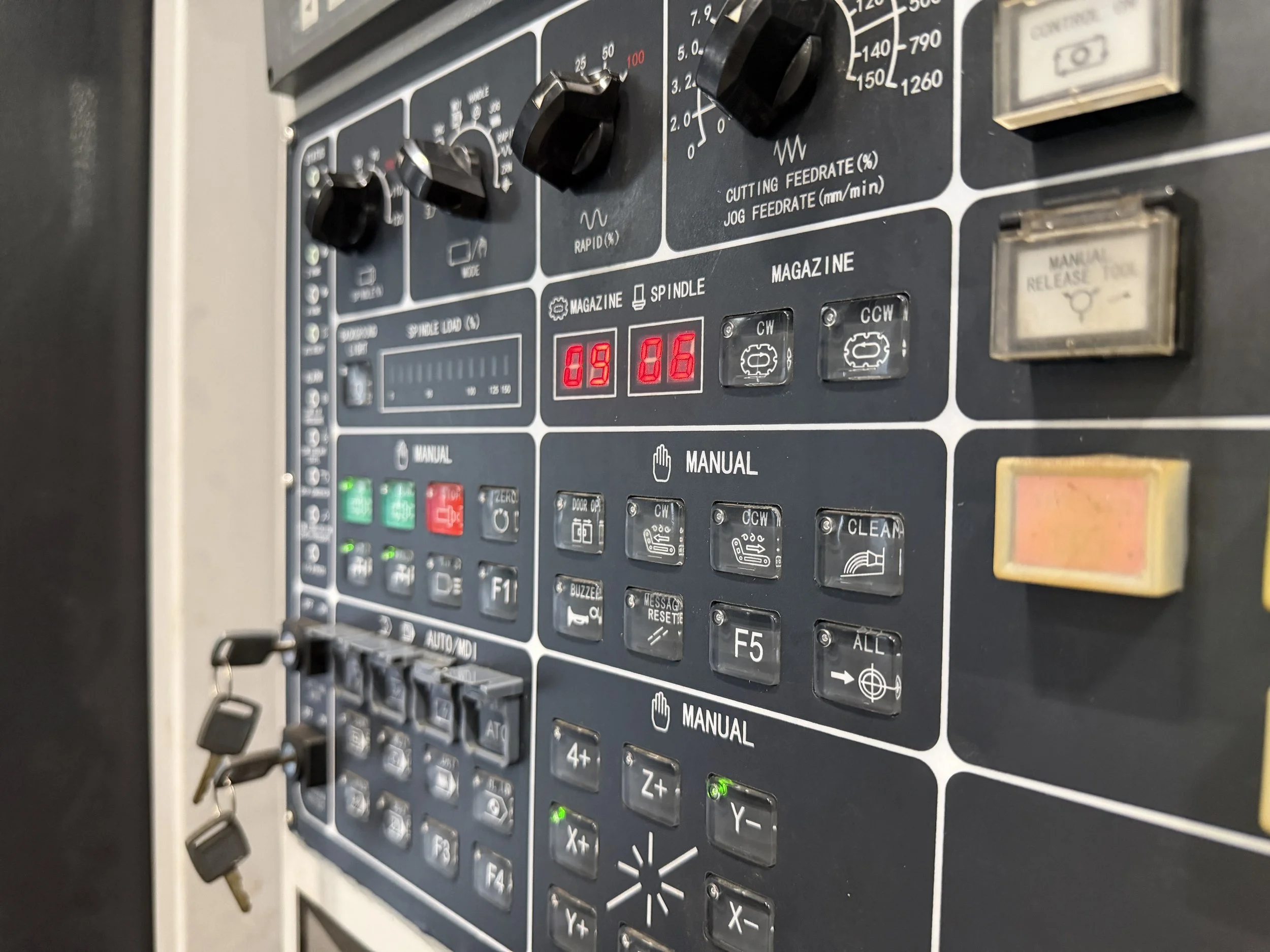 Close-up view of a CNC machine control panel with buttons, knobs, digital displays, and labels for various functions including spindle load, feed rates, and manual controls.