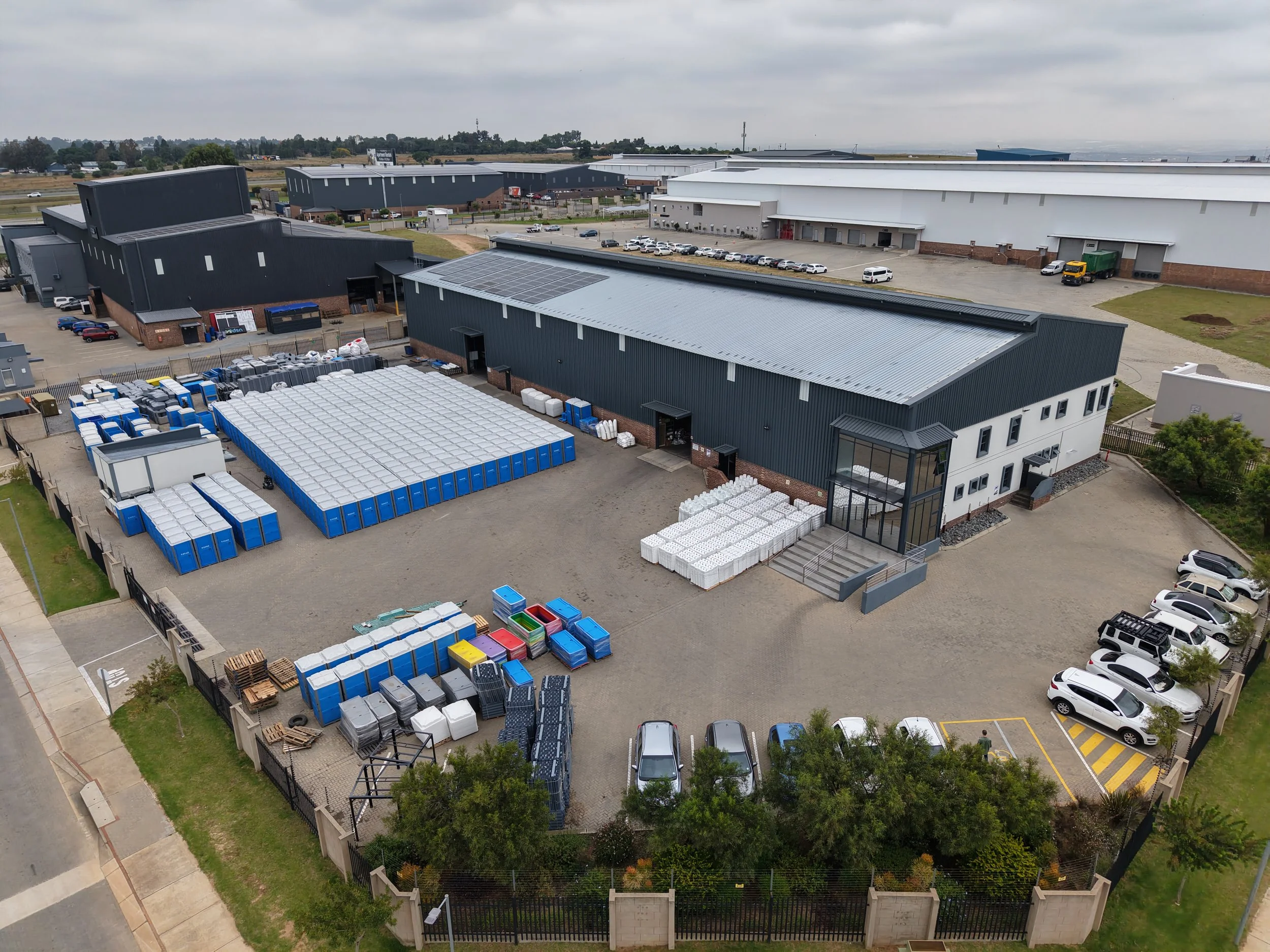 An aerial view of a commercial warehouse complex with parking lots, loading docks, and industrial storage containers, surrounded by a fence and some greenery.